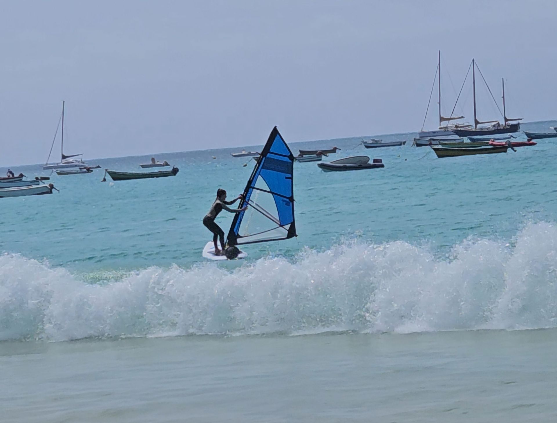Una donna che fa windsurf su acqua turchese con una grande onda in primo piano e diverse barche ancorate sullo sfondo.