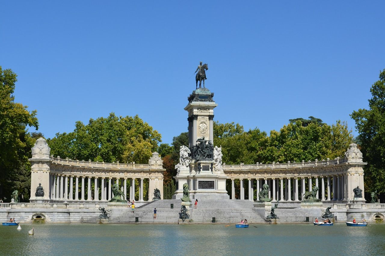 A large monument with an equestrian statue and a colonnade overlooks a lake with people rowing boats under a clear blue sky.