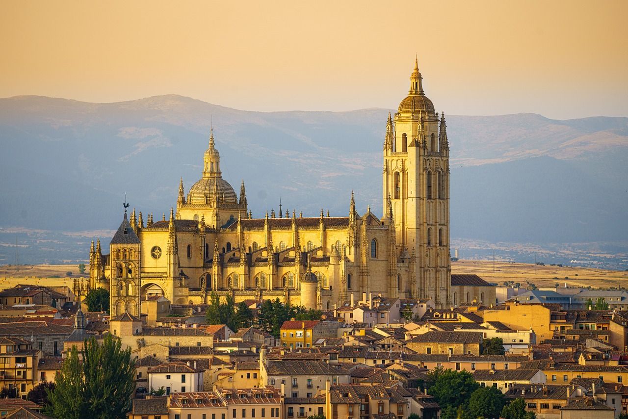 A grand Gothic cathedral with domes and a bell tower sits above a town at sunrise, with hazy mountains in the background.