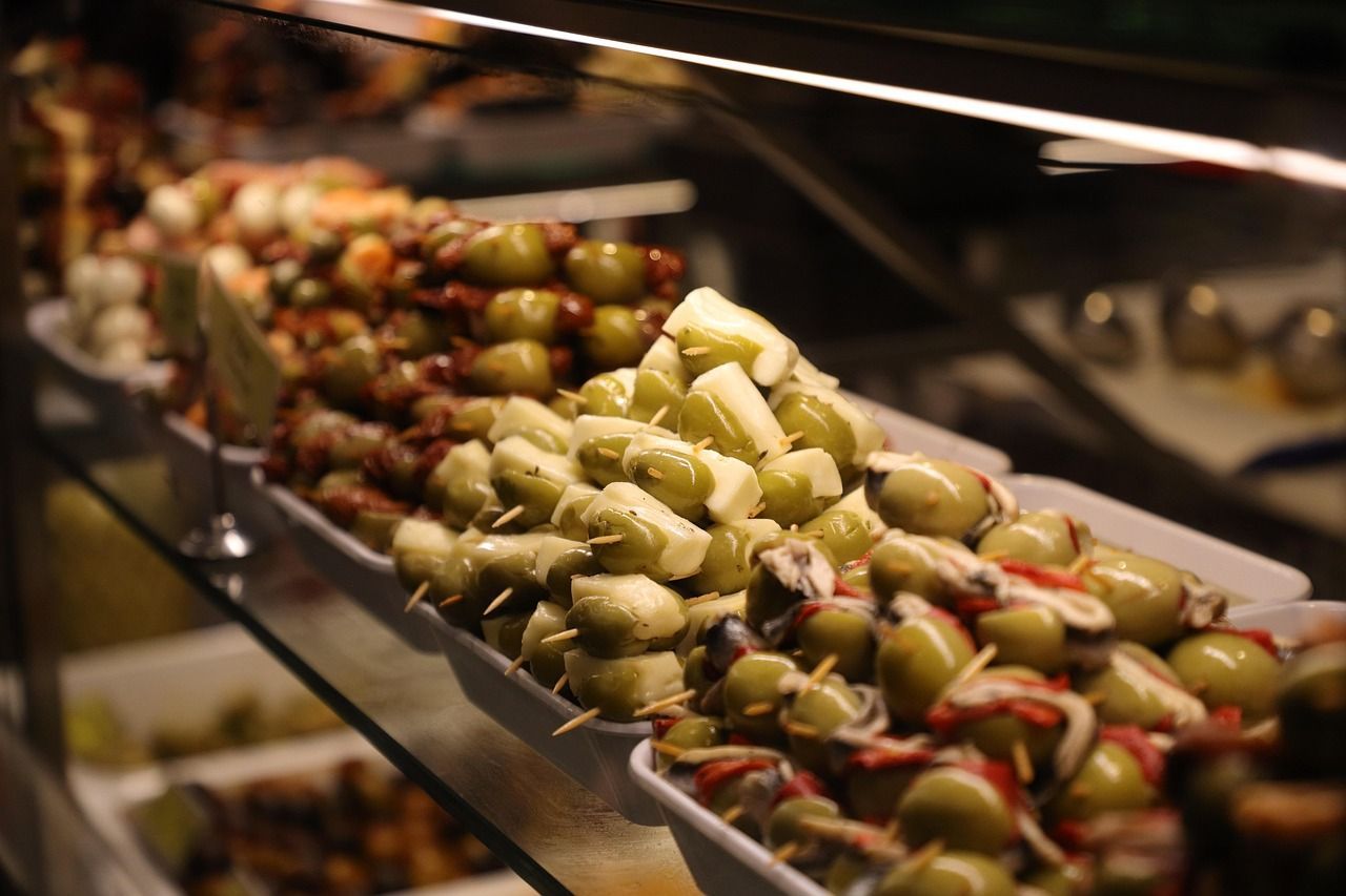 Trays of tapas skewers with green olives, cheese, and anchovies sit on a glass shelf in a display case.
