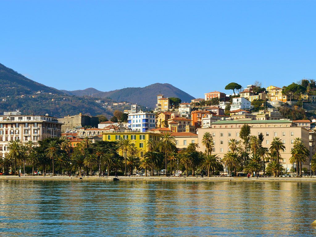 Una città sul lungomare con edifici colorati e palme si trova ai piedi di verdi colline sotto un cielo azzurro e limpido.