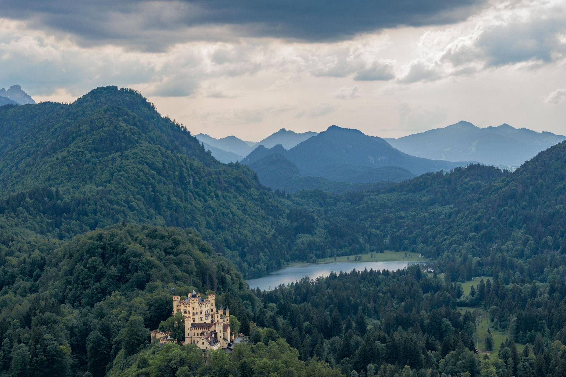 Un castello giallo si erge su una collina boscosa, con un lago e strati di montagne sullo sfondo sotto un cielo nuvoloso.