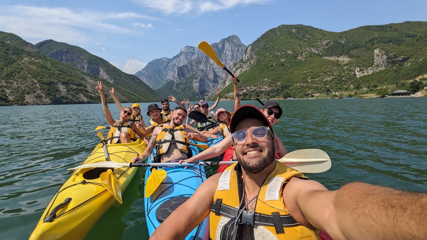 Un gruppo WeRoad si scatta un selfie sorridente in kayak su un lago con montagne verdi sullo sfondo.