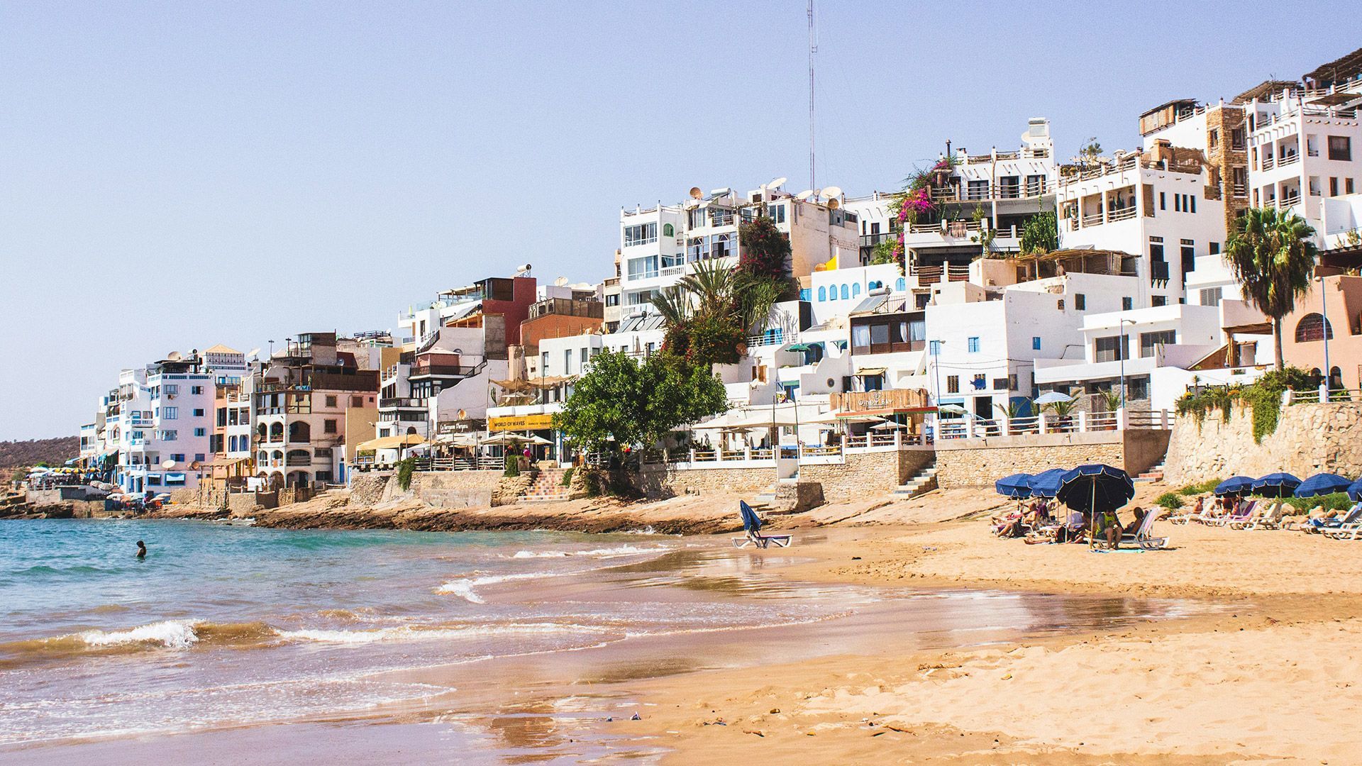 Une ville côtière aux bâtiments blancs surplombe une plage de sable où les gens se détendent sous des parasols bleus.