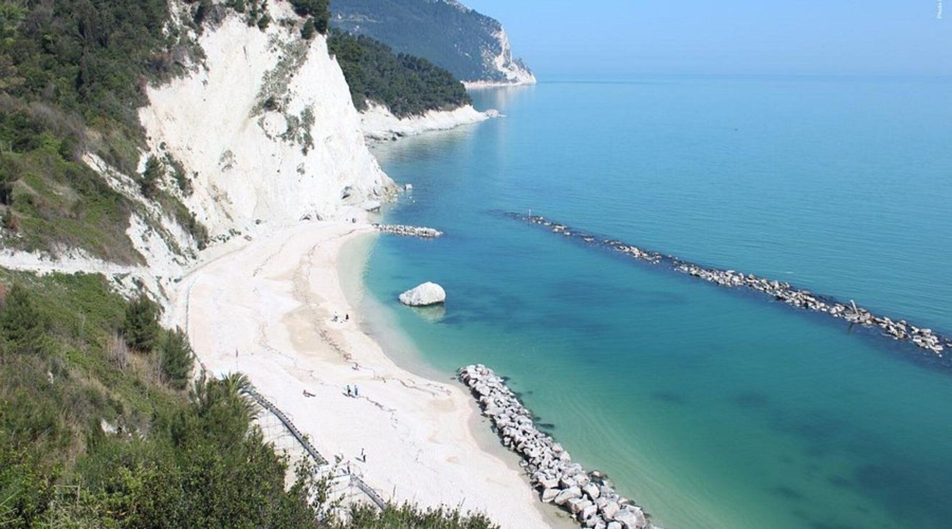 Una vista aerea di una spiaggia sabbiosa e appartata con acqua turchese, incastonata sotto una ripida scogliera bianca.