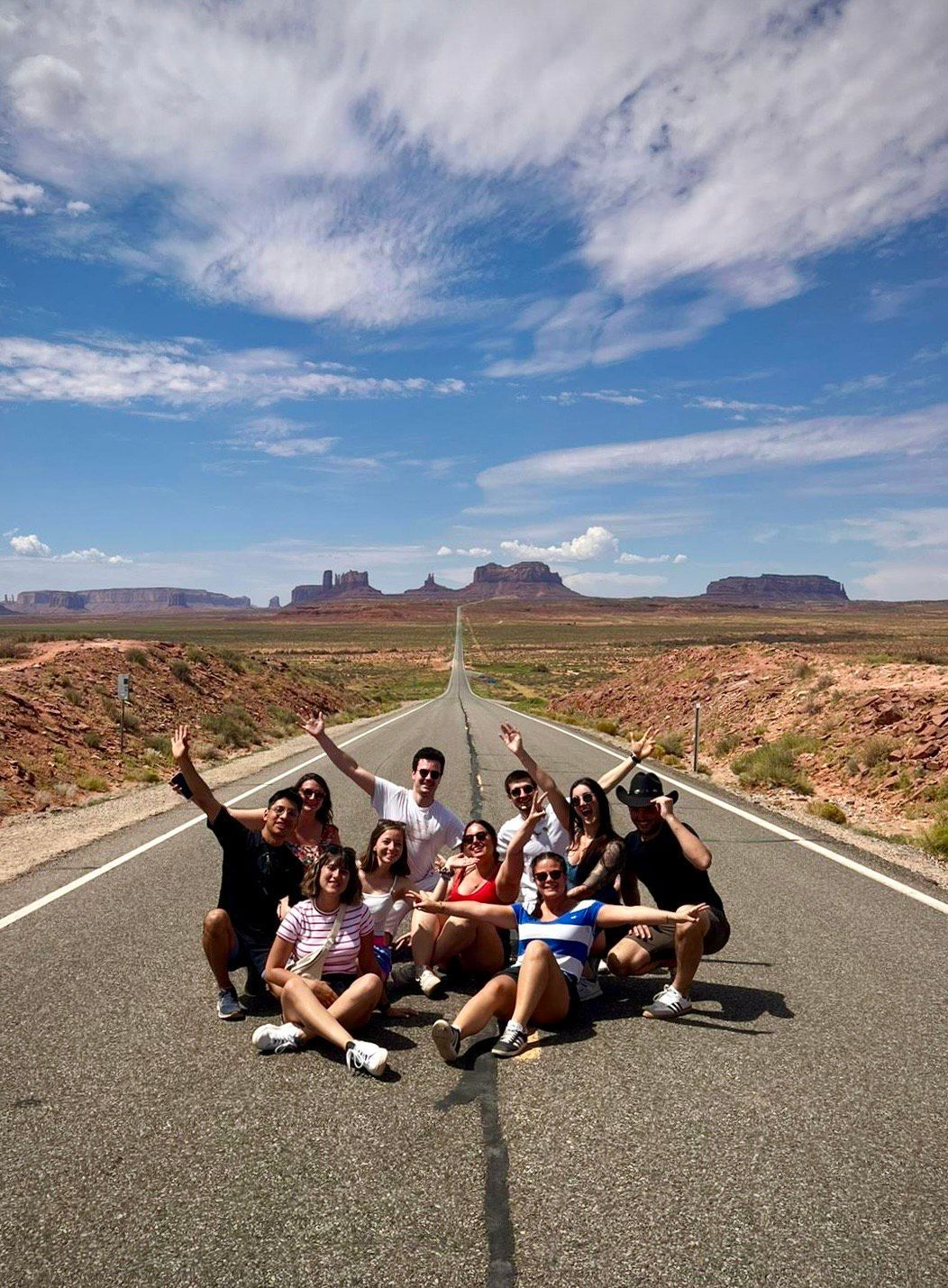 Un gruppo in viaggio con WeRoad posa al centro di una strada desertica che si estende verso lontane mesa sotto un cielo blu.