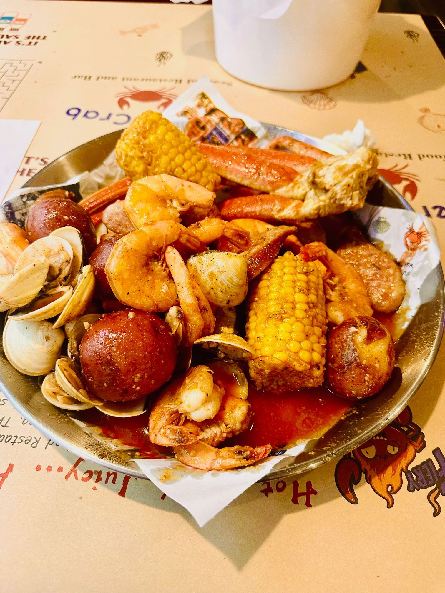 A close-up of a metal tray with a seafood boil of shrimp, crab legs, clams, corn, and potatoes covered in a red sauce.