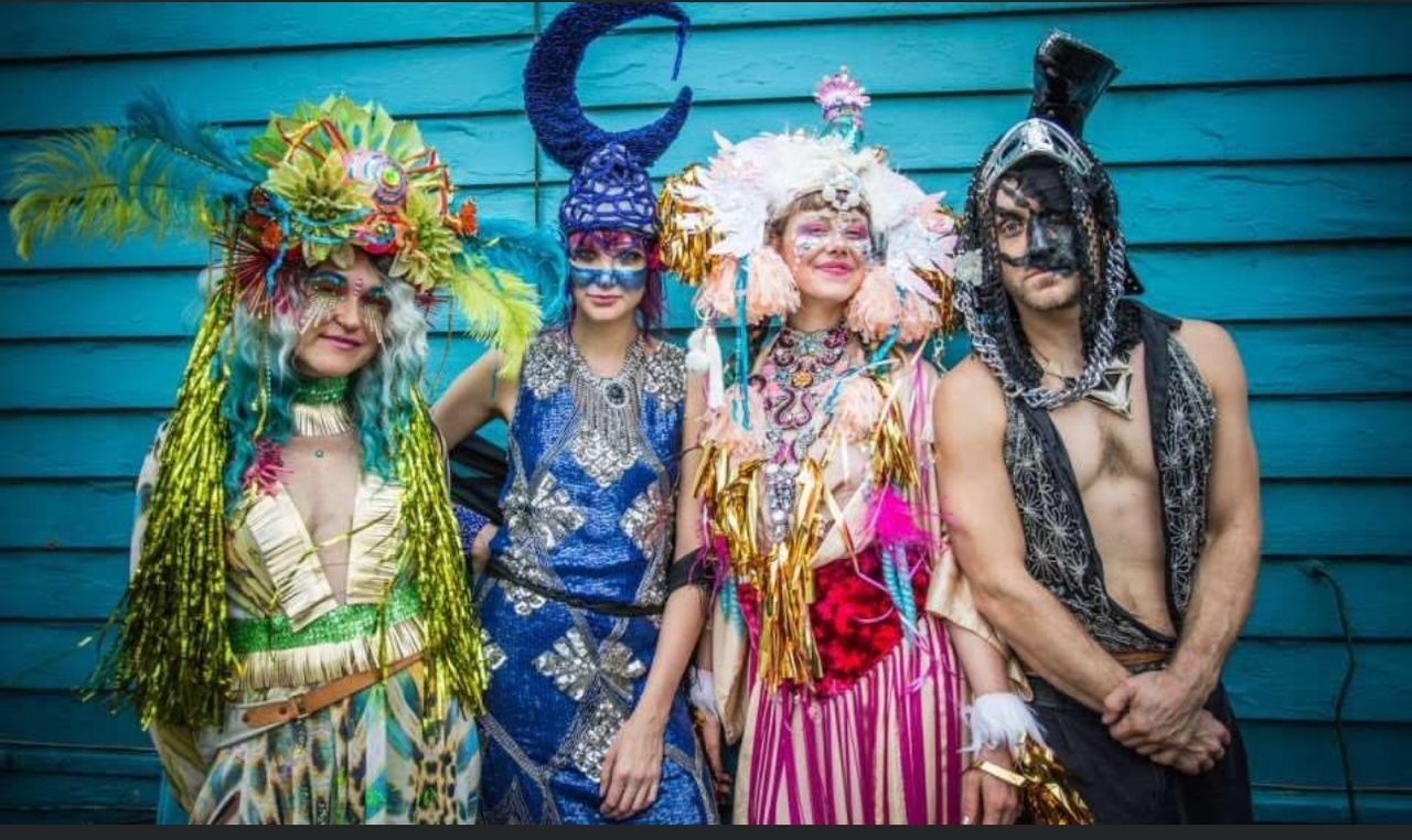 A WeRoad group trip of four people posing in elaborate, colorful costumes with headdresses and face paint against a blue wall.