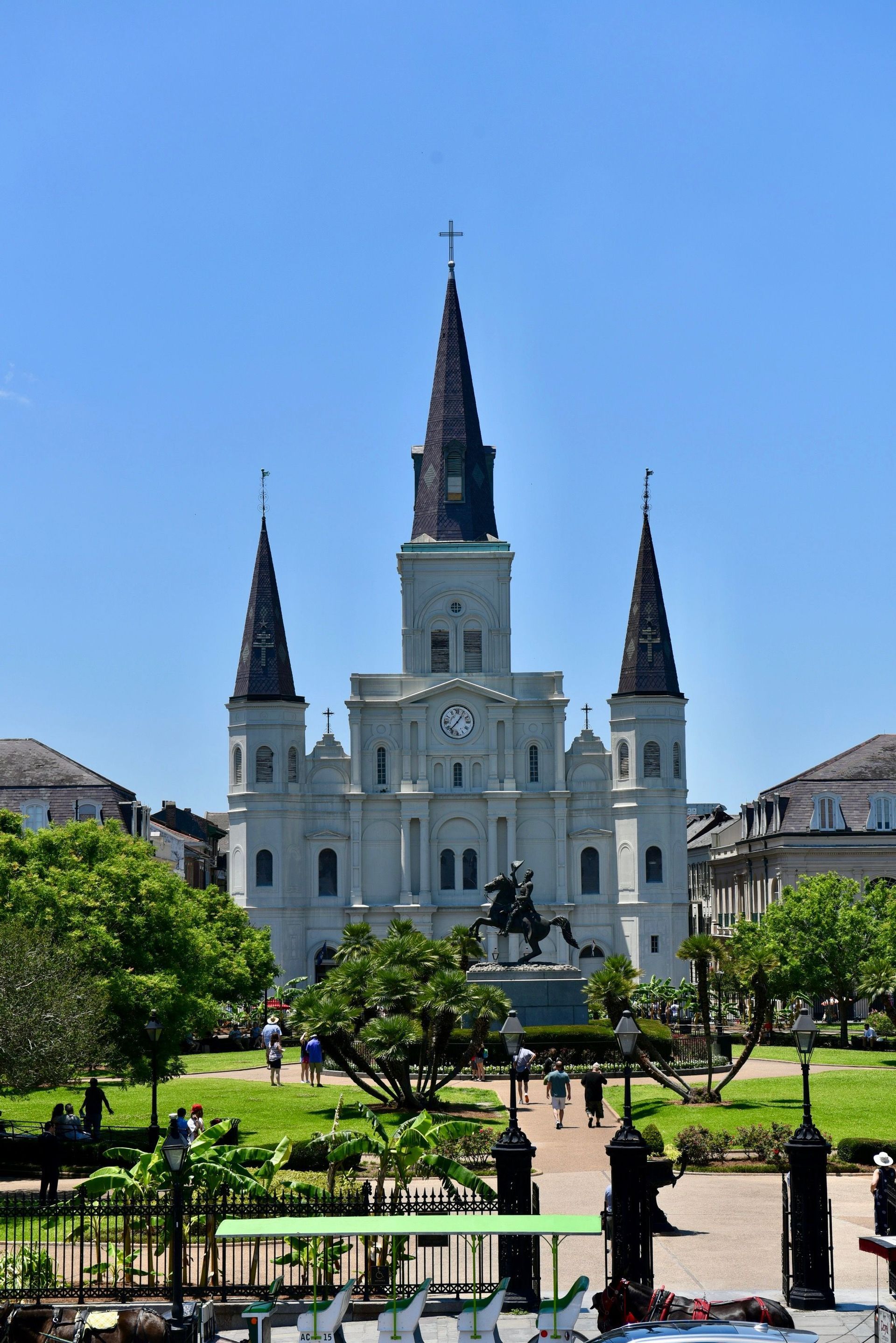 A large white cathedral with three steeples stands behind a green park containing an equestrian statue and people walking.