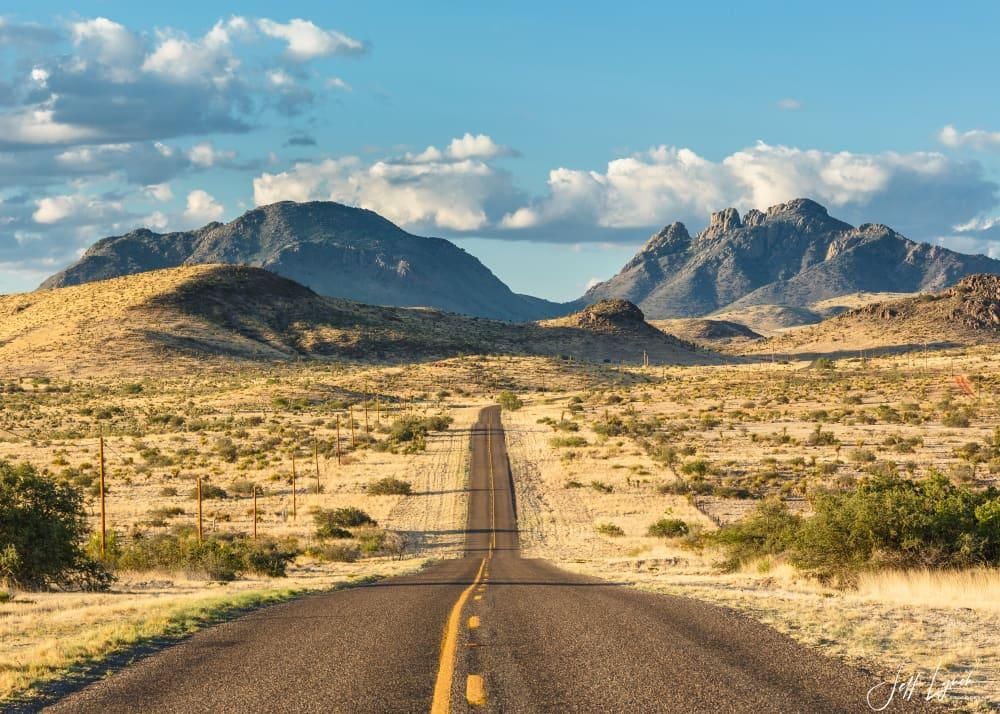 Una lunga strada asfaltata si estende attraverso un paesaggio desertico verso montagne lontane sotto un cielo azzurro nuvoloso.
