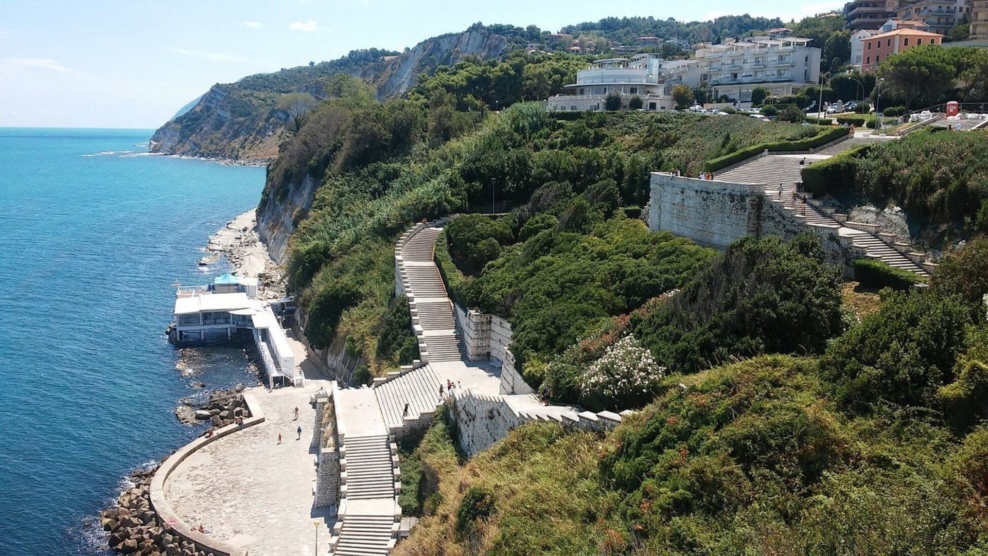 Vista aerea di un'ampia scalinata in pietra che si snoda lungo un rigoglioso pendio verde verso una costa rocciosa e il mare blu.