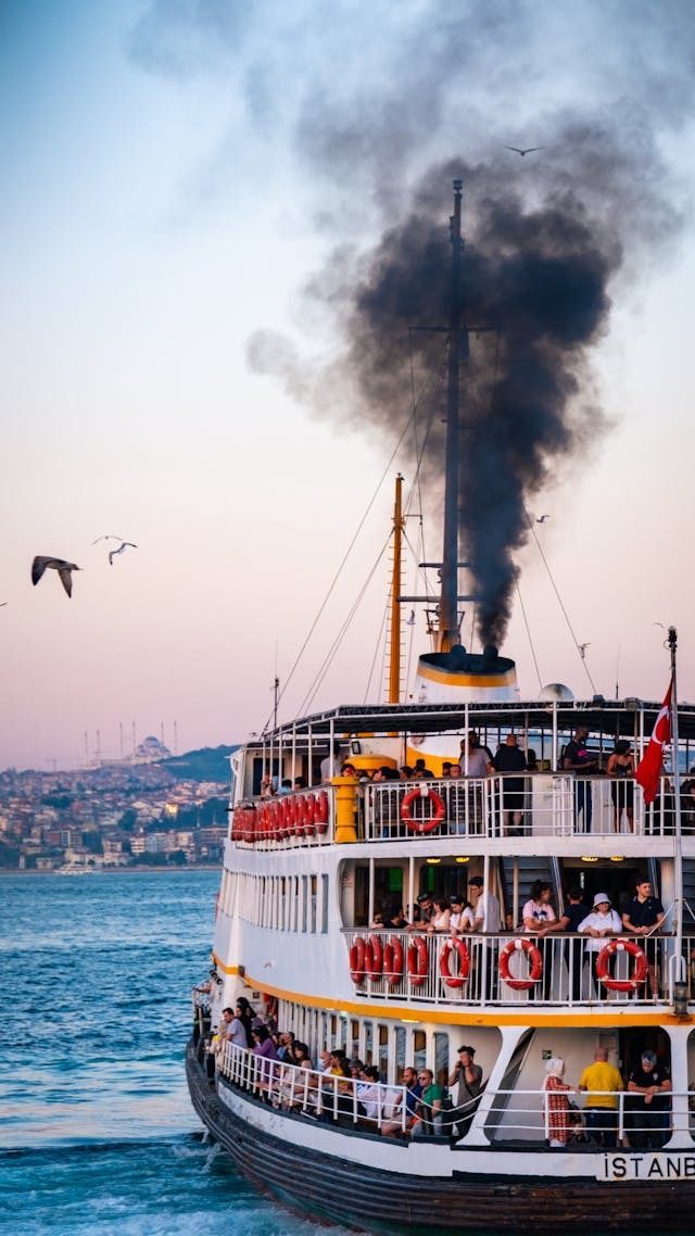 A multi-deck ferry with passengers sails on the water, releasing a thick plume of black smoke from its smokestack against a pale sky.