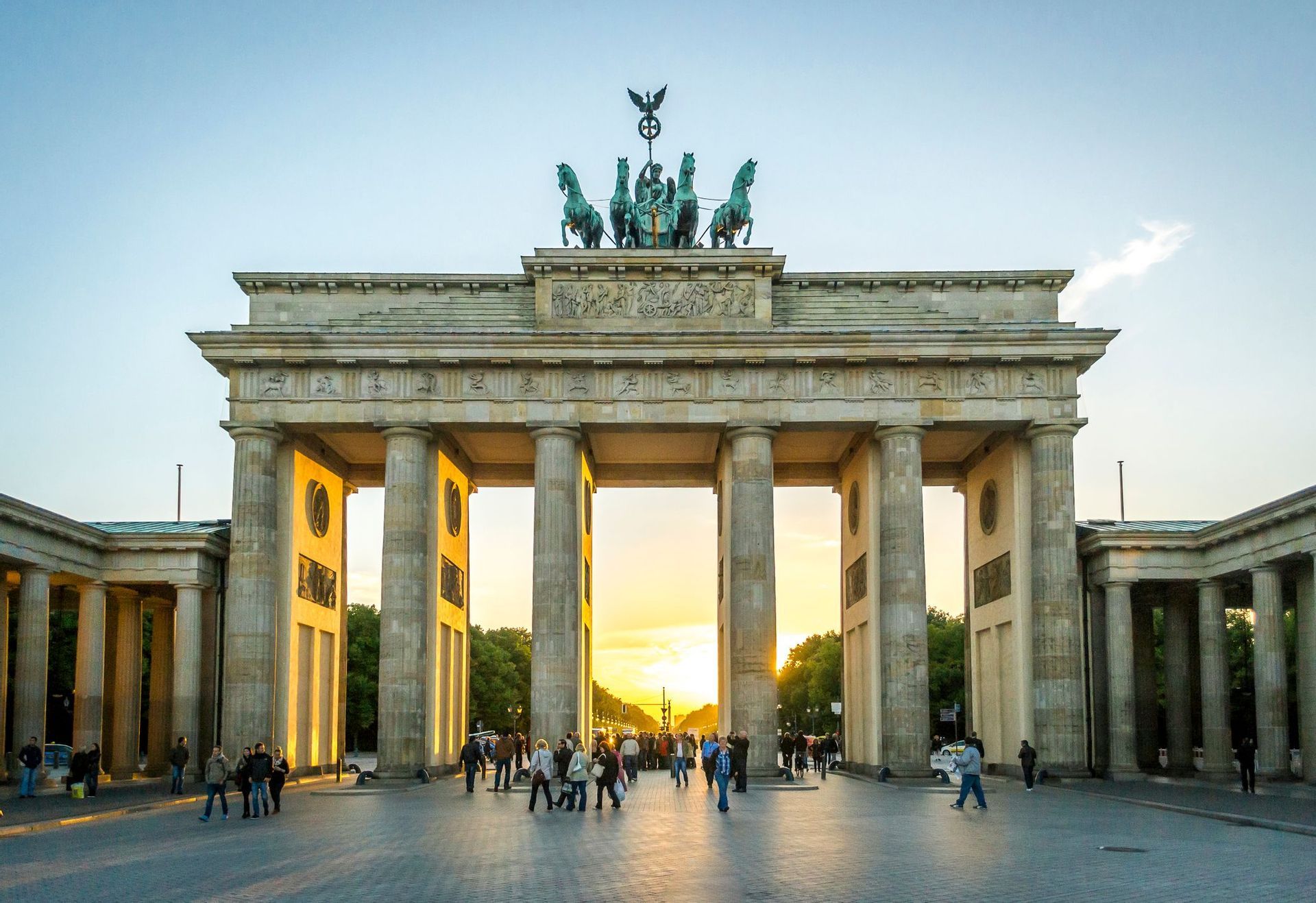A grand neoclassical monument with a quadriga statue on top, with people walking in the plaza below during sunset.