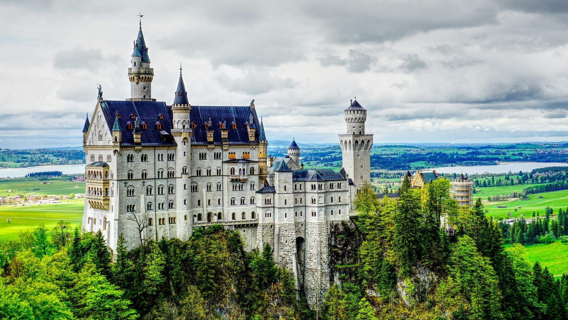 A large castle with white walls and blue roofs stands on a forested hill, overlooking a vast green valley and a lake under a cloudy sky.