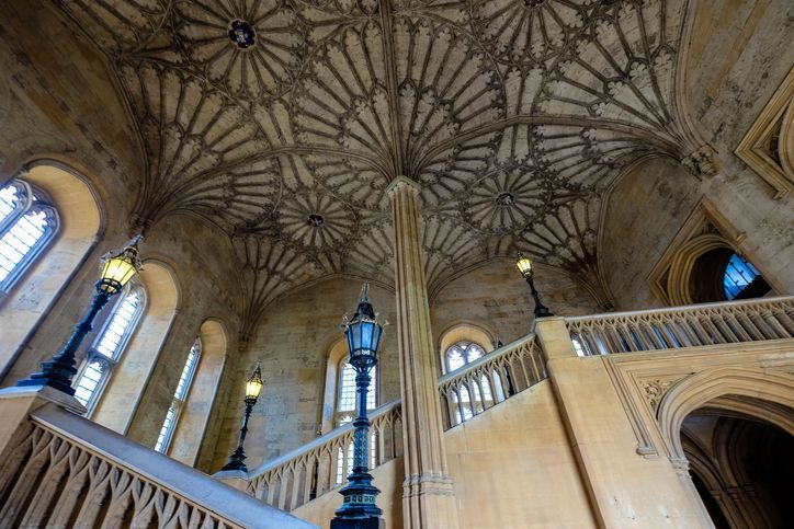 A low-angle view of a grand stone staircase with ornate lampposts leading up towards an intricate fan-vaulted ceiling.