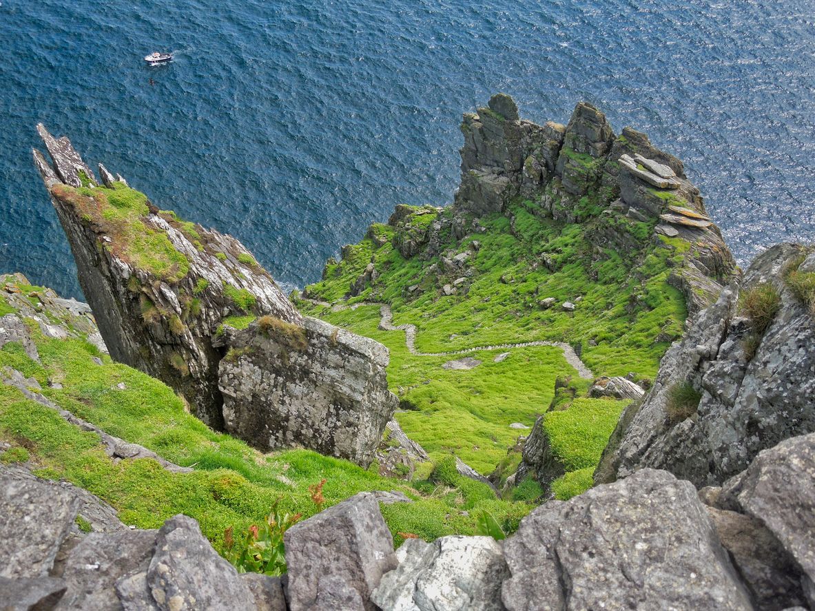 A high-angle view of a steep, grassy cliff with a winding stone path leading down towards a deep blue ocean with a boat.