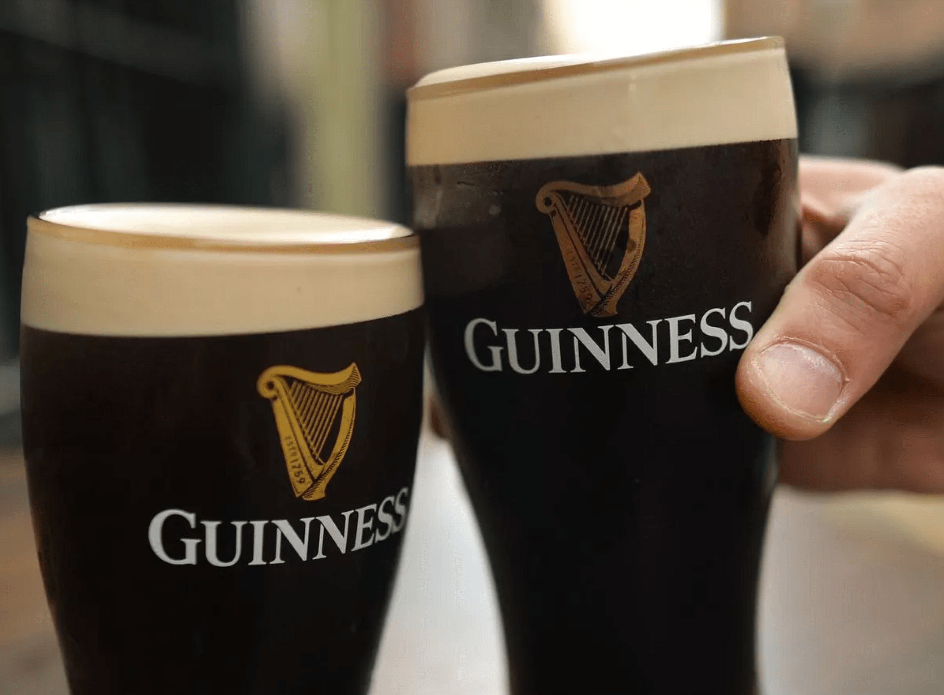 A close-up of two pint glasses of Guinness stout with creamy heads, with a hand holding one of the glasses.