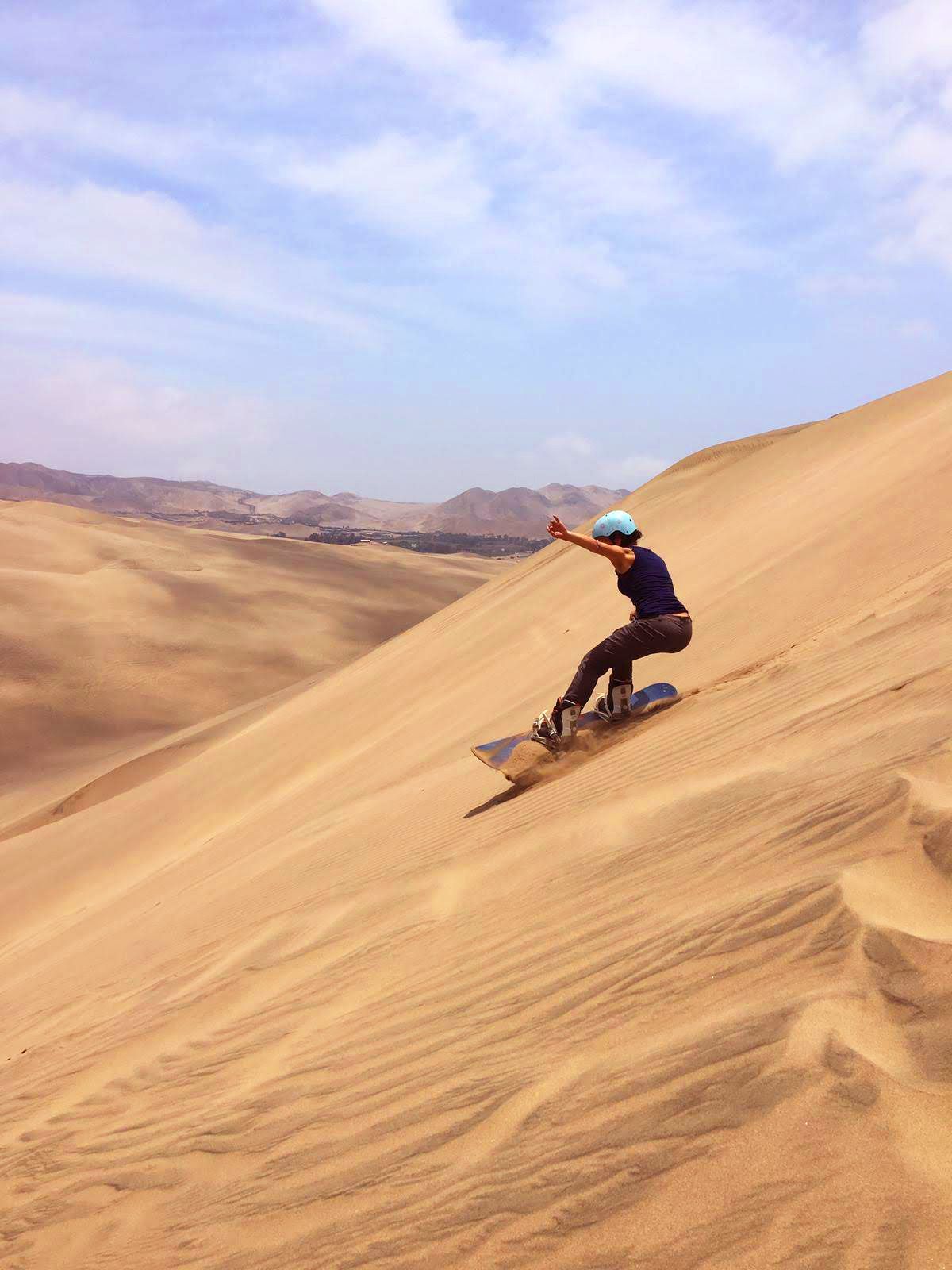 Une personne portant un casque fait du sandboard sur une grande et raide dune de sable dans un vaste désert sous un ciel partiellement nuageux.