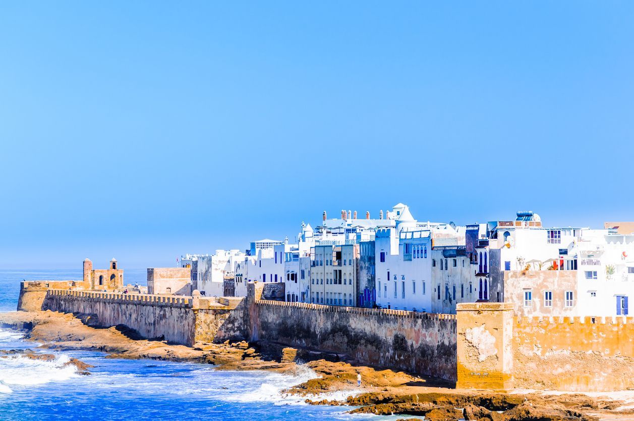 A fortified coastal city with whitewashed buildings stands on a rocky shore, with blue ocean waves in the foreground under a clear sky.