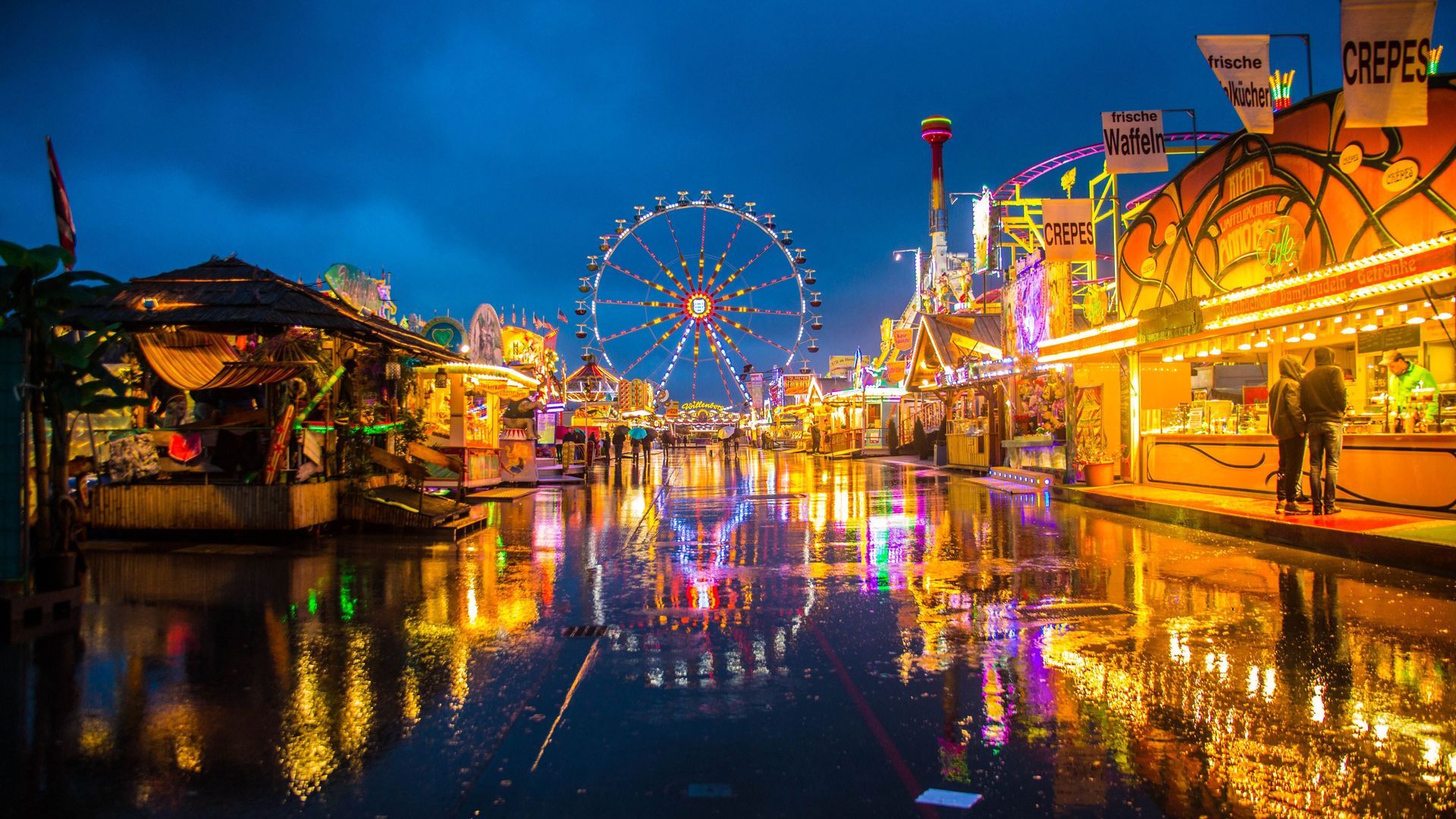 Un parque de atracciones iluminado al anochecer con una noria y puestos de comida, sus luces de colores reflejándose en la calle mojada.