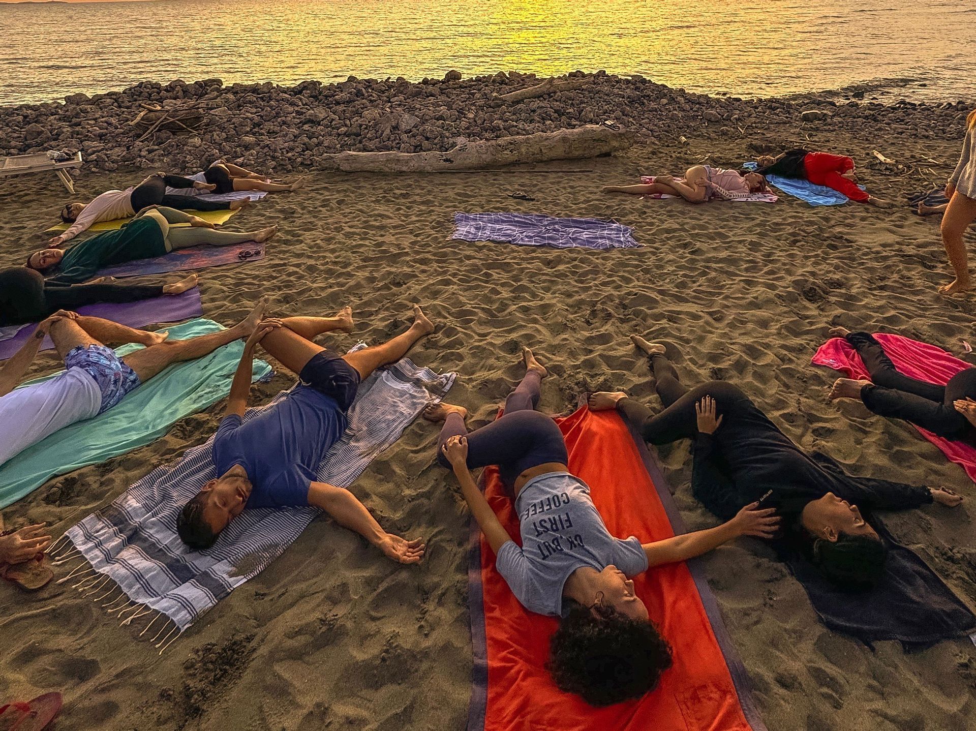 Un gruppo WeRoad in viaggio si rilassa sugli asciugamani su una spiaggia sabbiosa, facendo stretching al tramonto.