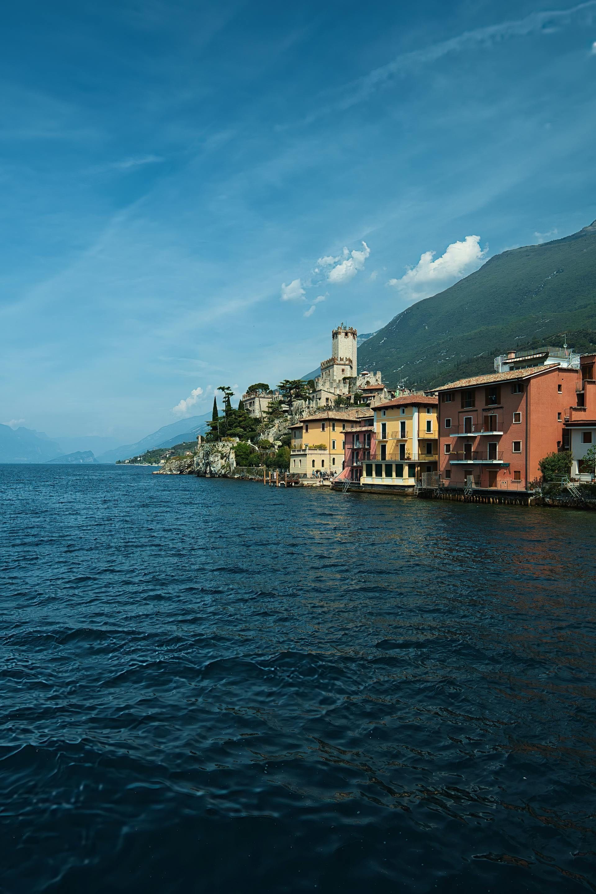 A lakeside village with colorful houses and a castle on a cliff, seen from the water with a mountain behind.