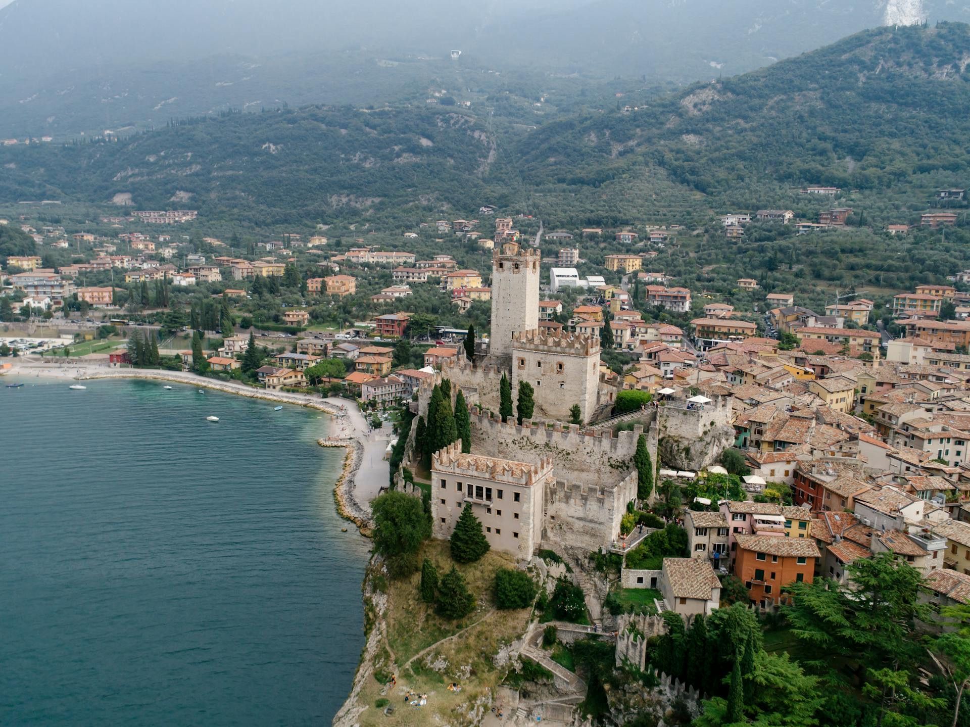 An aerial view of a stone castle on a cliff overlooking a lakeside town, with green mountains in the background.