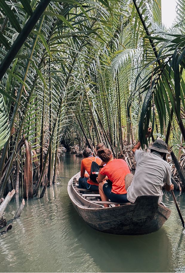 A WeRoad group trip travels by wooden boat through a narrow canal lined with dense, tall palm trees.
