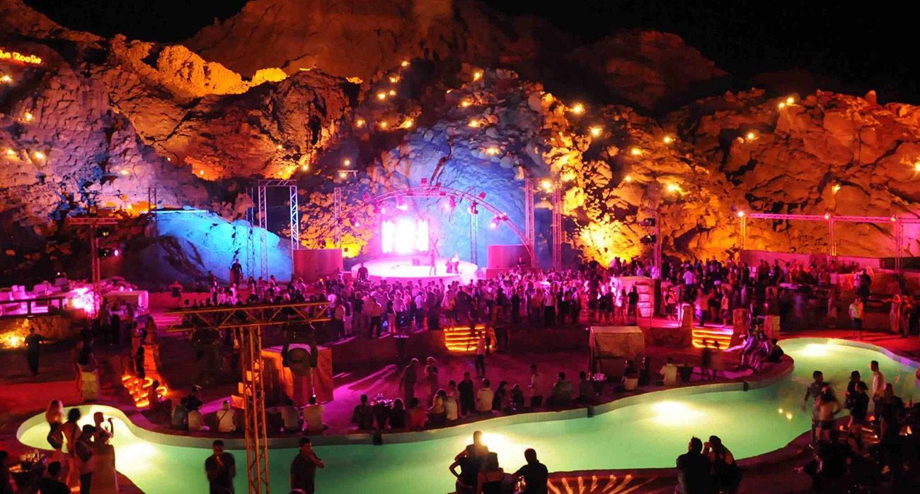 Una grande folla di persone a un evento notturno con piscina e palco, sullo sfondo di formazioni rocciose illuminate drammaticamente.