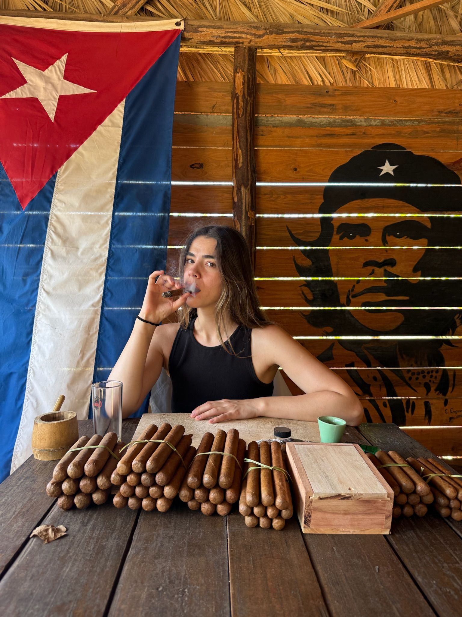 Une femme fume un cigare à une table couverte de paquets de cigares, avec un drapeau cubain et une fresque de Che Guevara en arrière-plan.