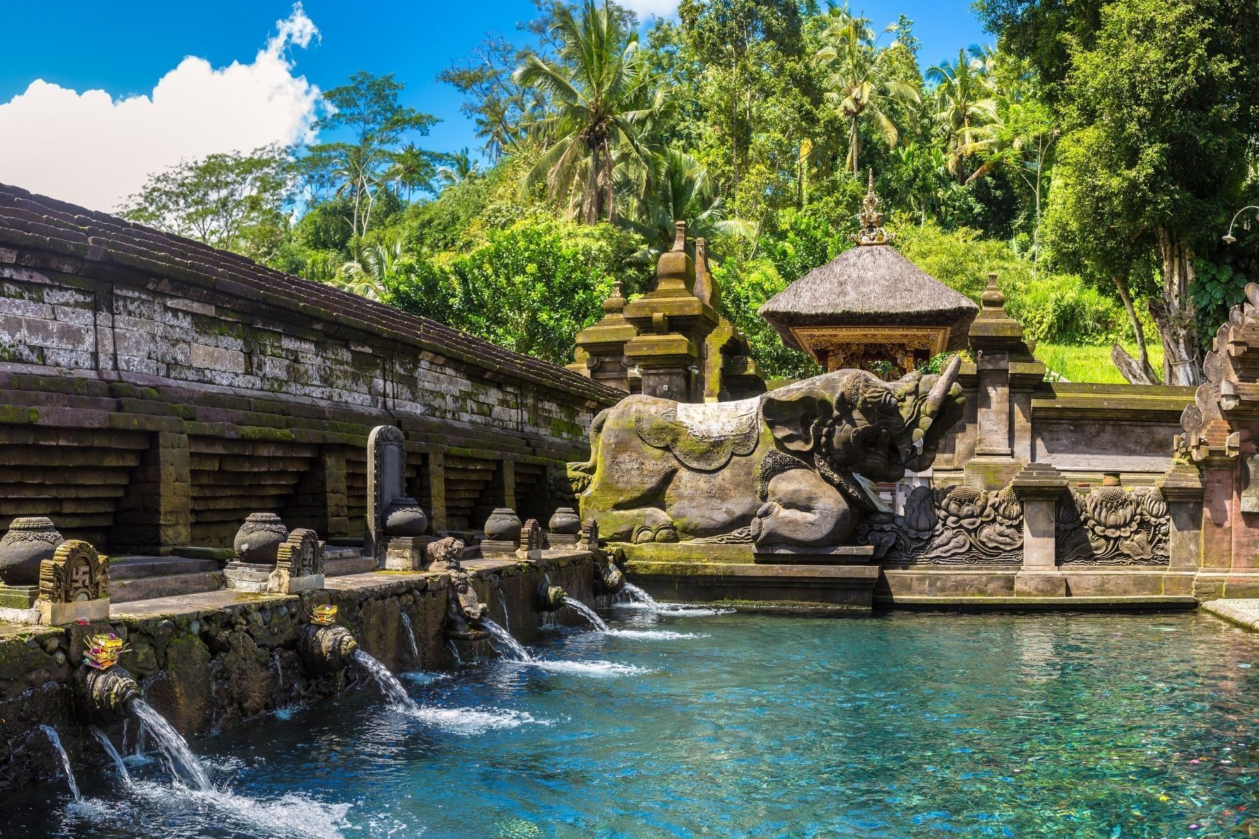 Steinerne Brunnen speien Wasser in einen klaren blauen Pool an einem moosbewachsenen Tempel mit einer großen Elefantenstatue und Dschungelkulisse.