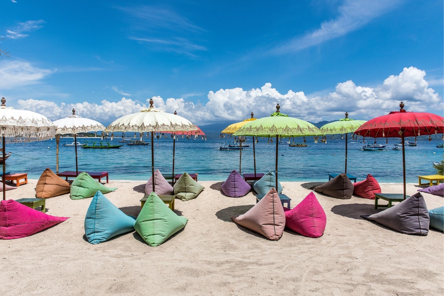 Bunte Sitzsäcke und traditionelle Sonnenschirme säumen einen Sandstrand vor einem türkisfarbenen Meer mit Booten unter blauem Himmel.