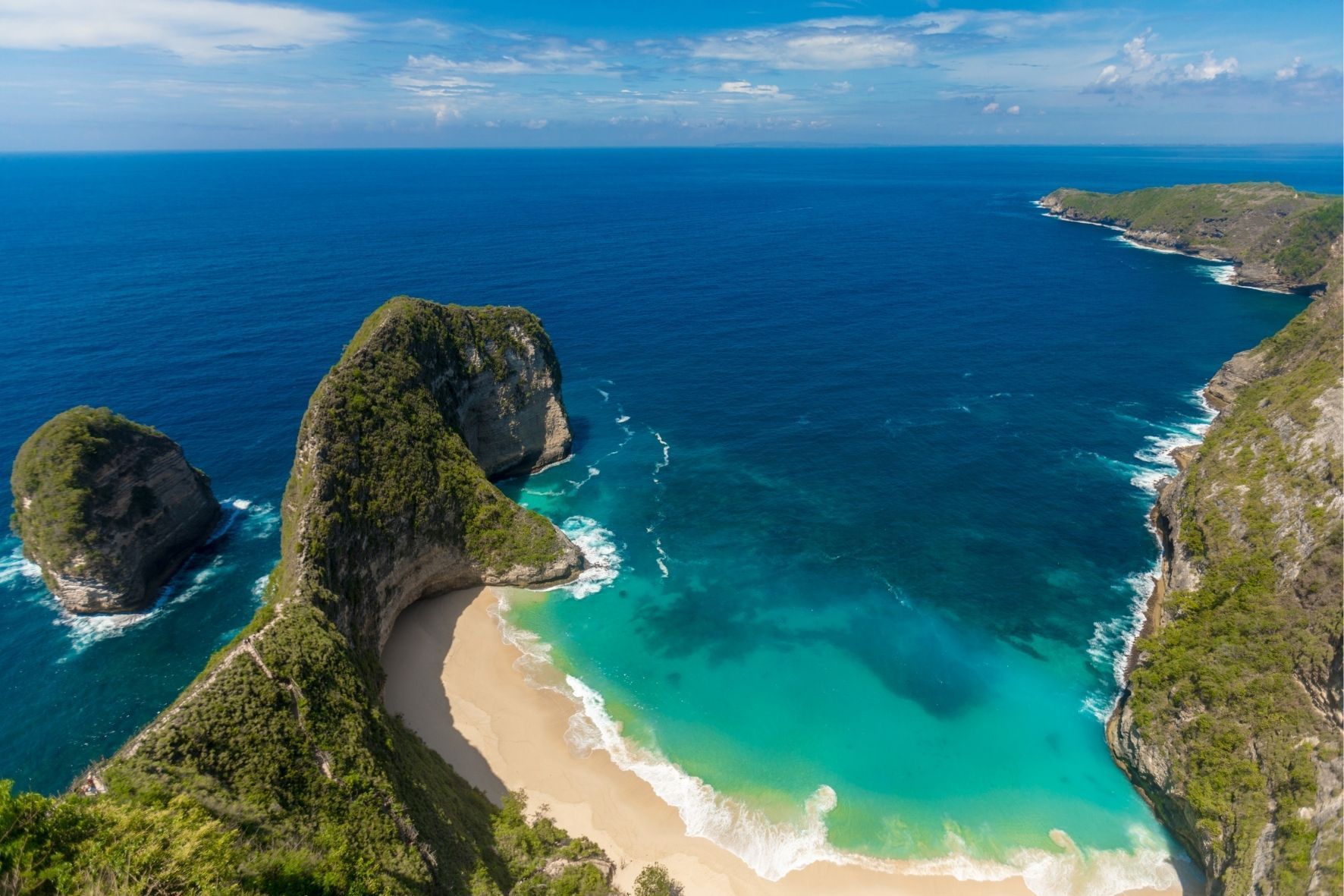 Eine Luftaufnahme einer großen, bewachsenen Felsformation neben einem weißen Sandstrand mit türkisfarbenem Wasser.