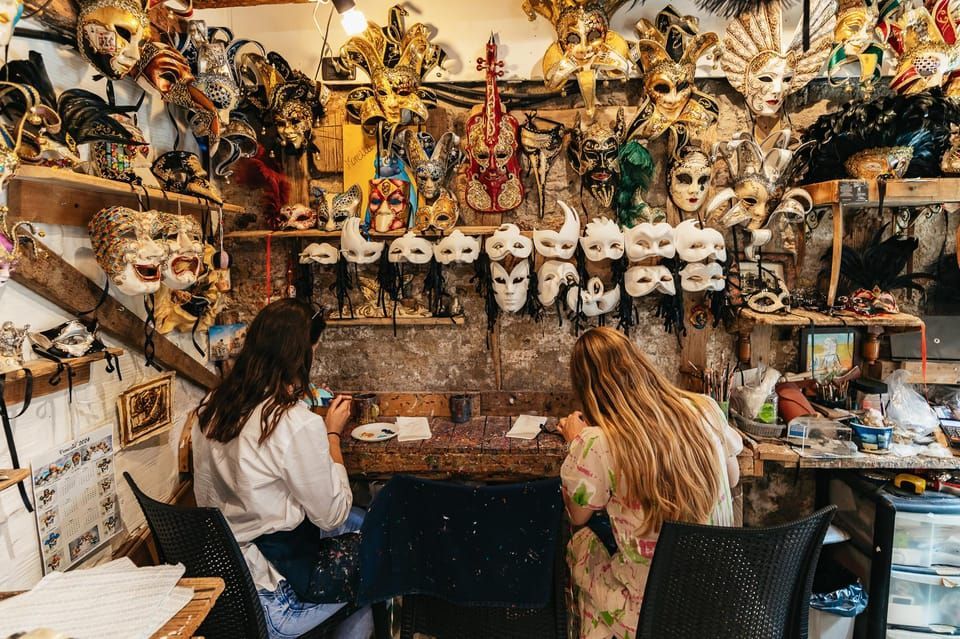 Deux femmes participant à un voyage de groupe WeRoad peignent des masques à une table dans un atelier rempli de masques vénitiens.