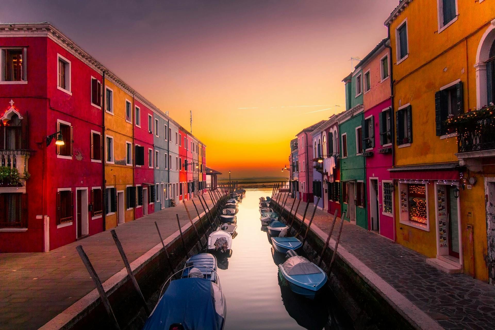 Small boats are moored in a canal between rows of colorful buildings under a glowing sunset sky.