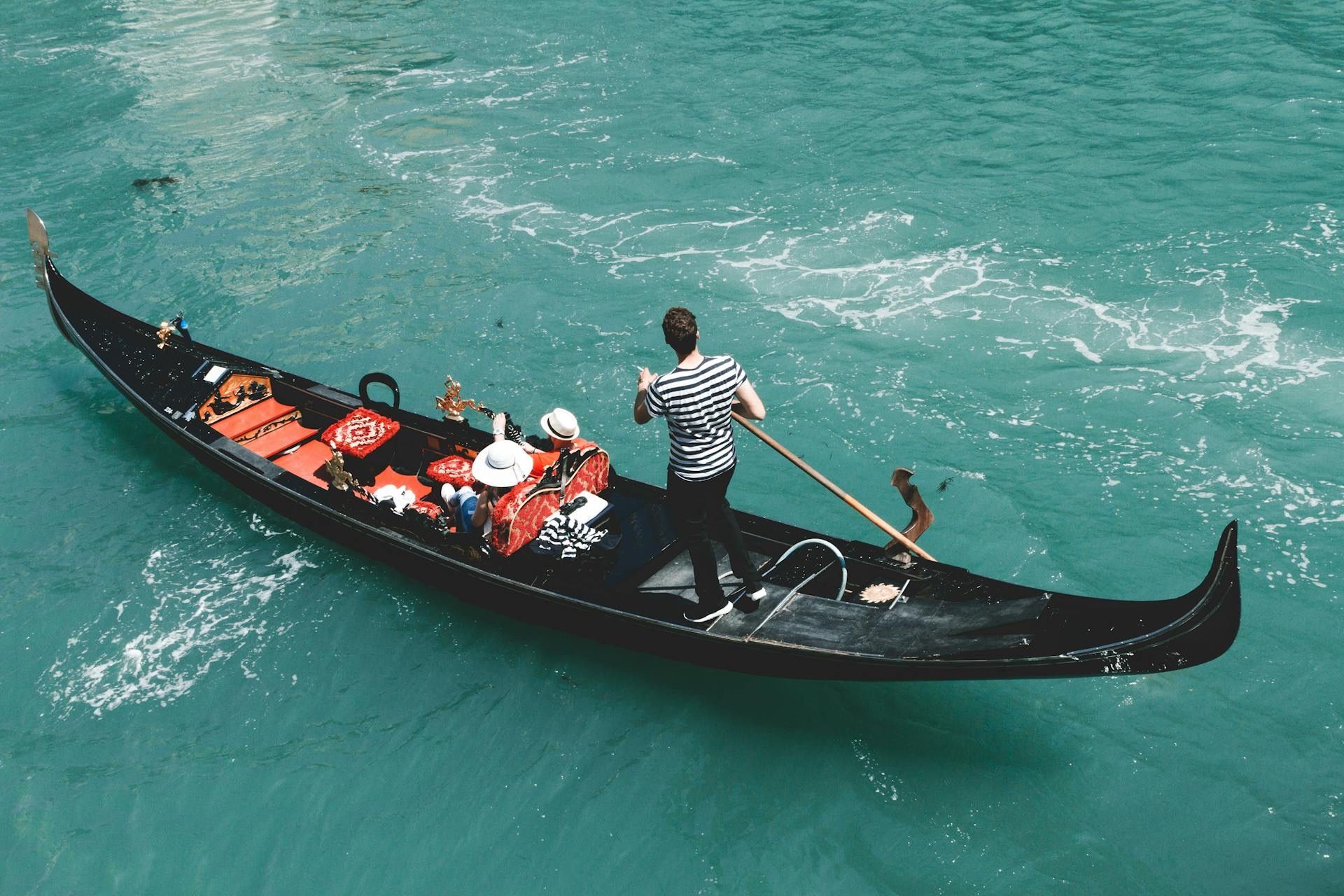 A gondolier in a striped shirt rows a gondola with two passengers from a WeRoad group trip across turquoise water.