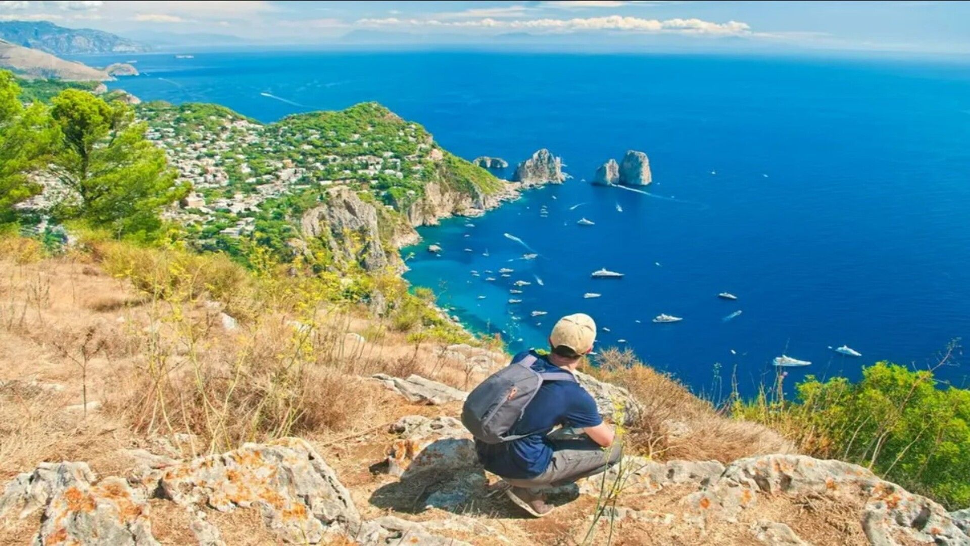 Una persona con mochila se agacha en lo alto de un acantilado rocoso, contemplando un mar azul intenso con barcos, farallones y un pueblo costero.