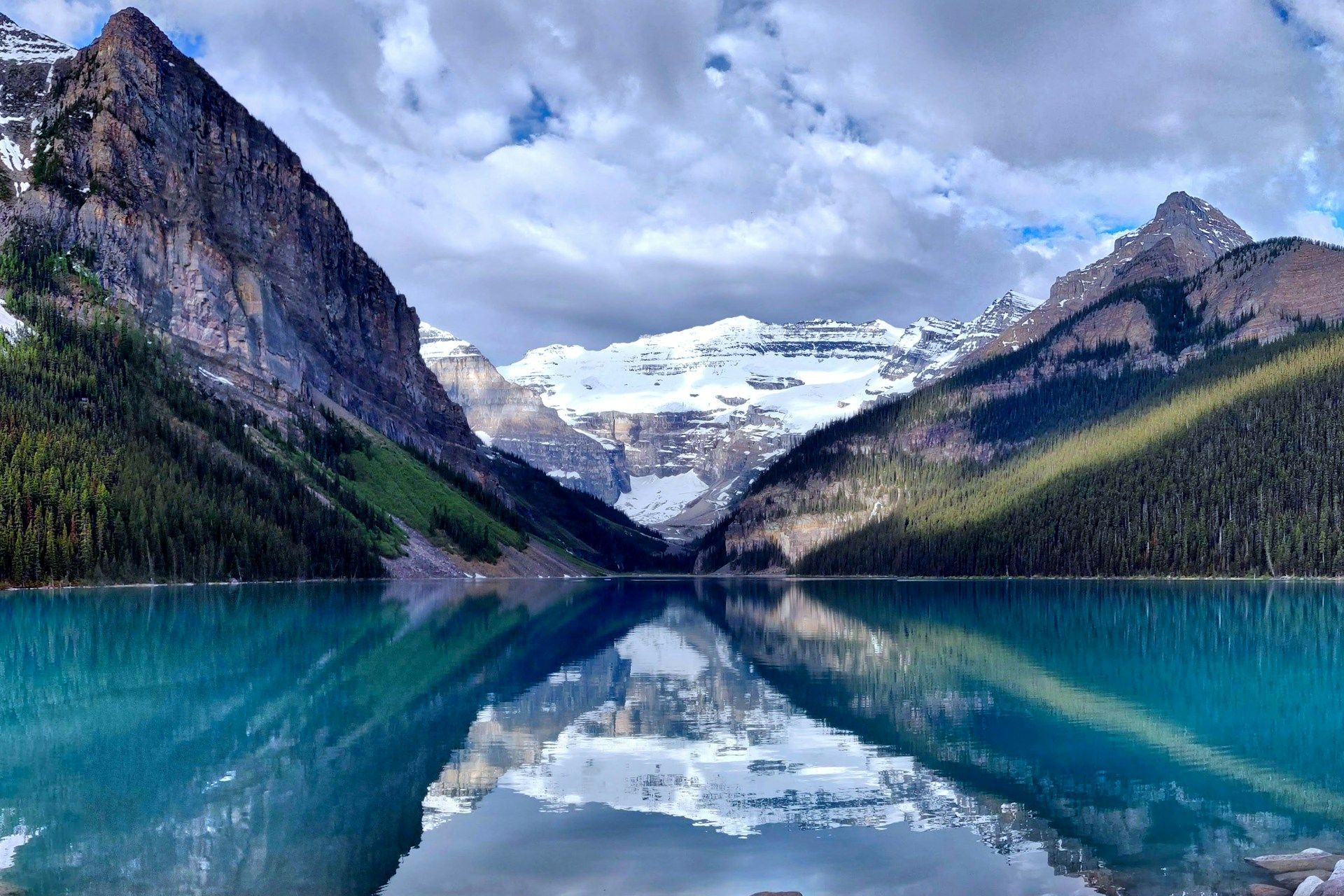 Snow-capped mountains and pine forests are reflected in the calm, turquoise water of an alpine lake under a cloudy sky.