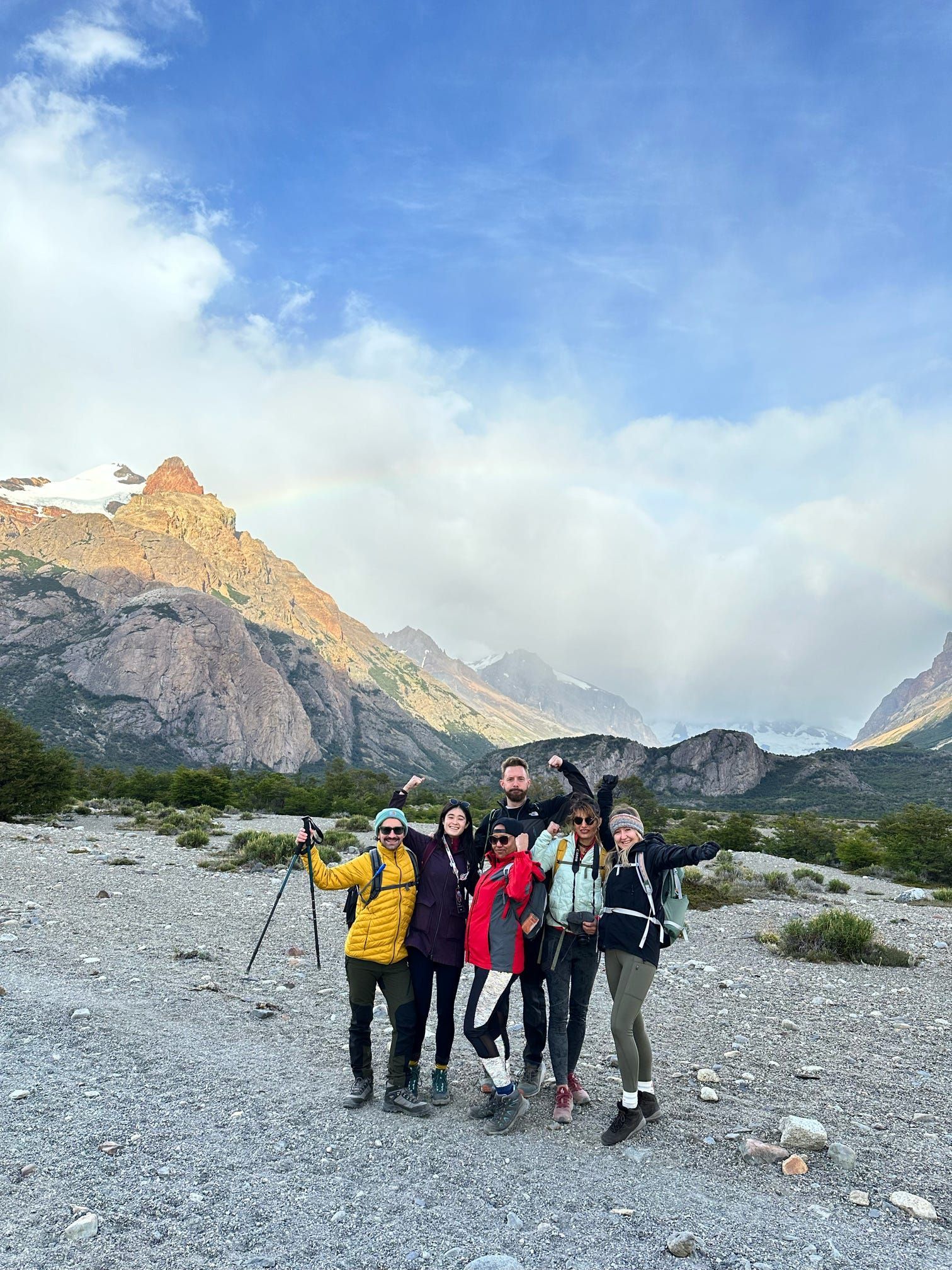 Un viaggio di gruppo WeRoad con attrezzatura da trekking posa in una valle montuosa rocciosa con un debole arcobaleno sullo sfondo.