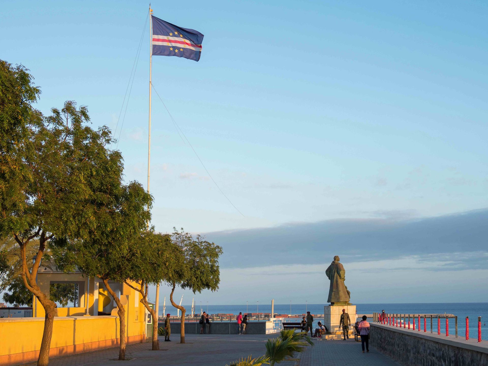 Le drapeau du Cap-Vert flotte sur une promenade en bord de mer, où une statue surplombe l'océan, à côté d'un bâtiment jaune.
