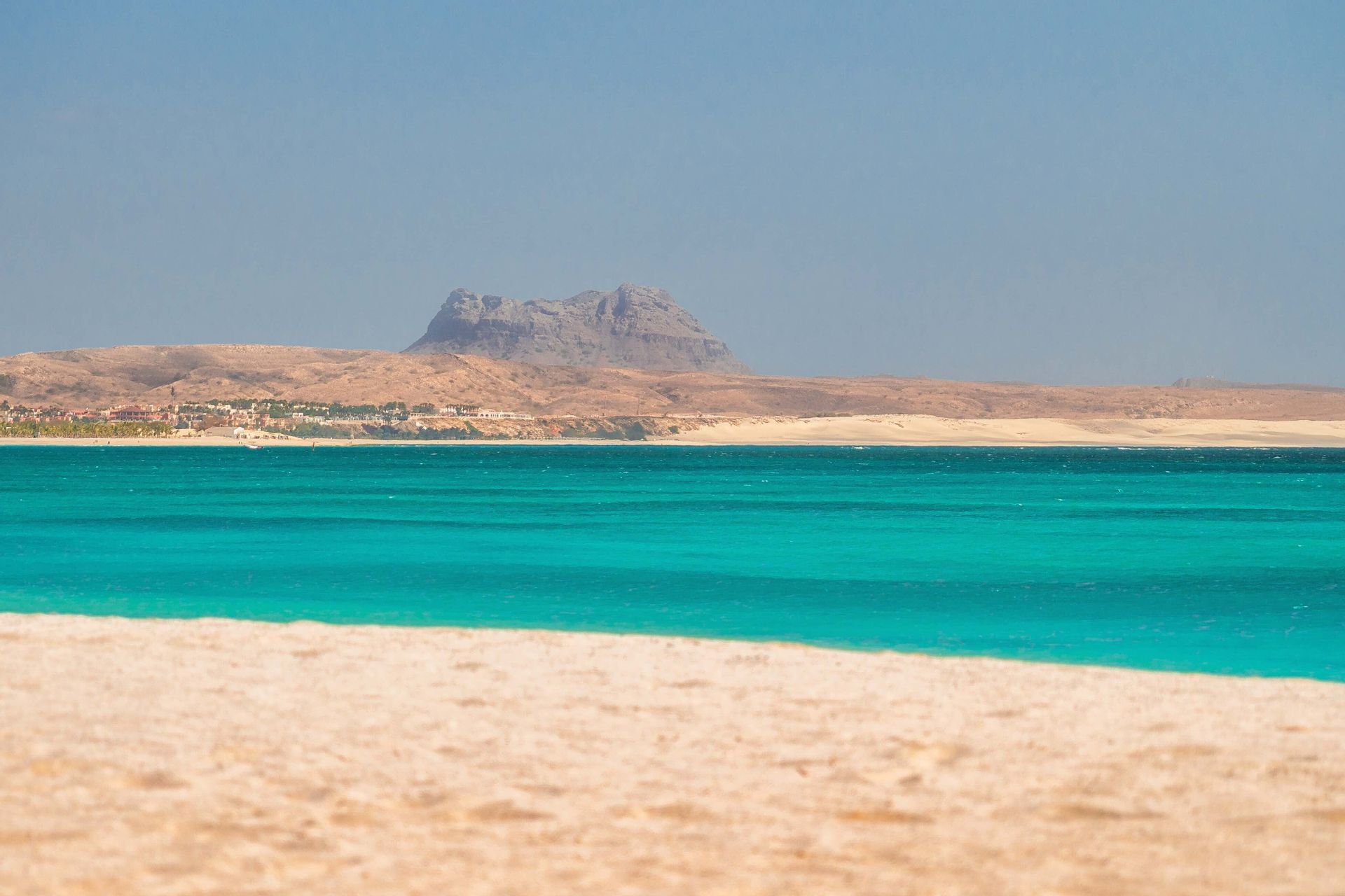 Une vue depuis une plage de sable fin, traversant une eau turquoise jusqu'à un paysage aride lointain avec une montagne sous un ciel bleu clair.