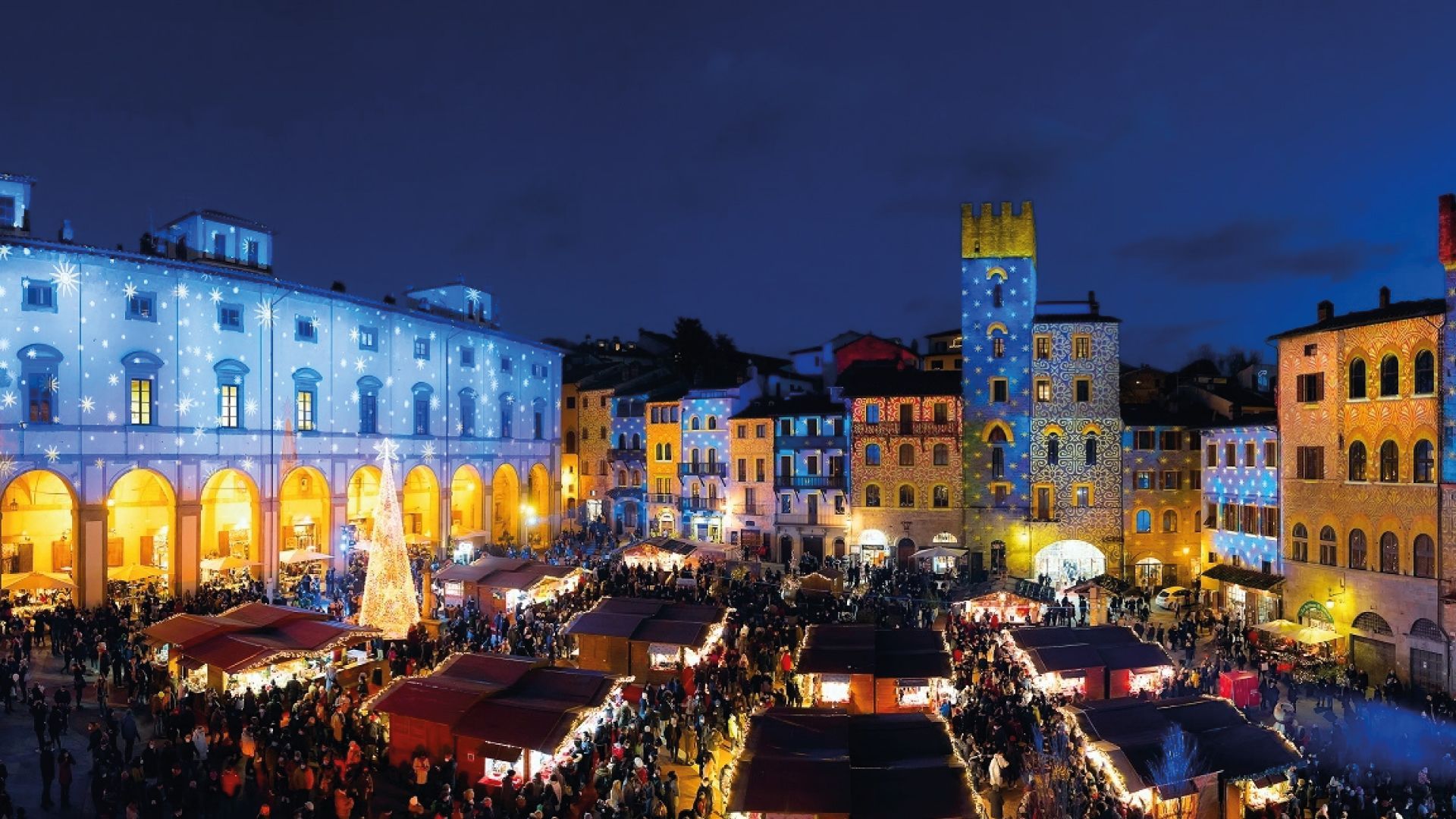Vista aerea di un affollato mercatino di Natale in una piazza cittadina di notte, con edifici illuminati da proiezioni di luci colorate.