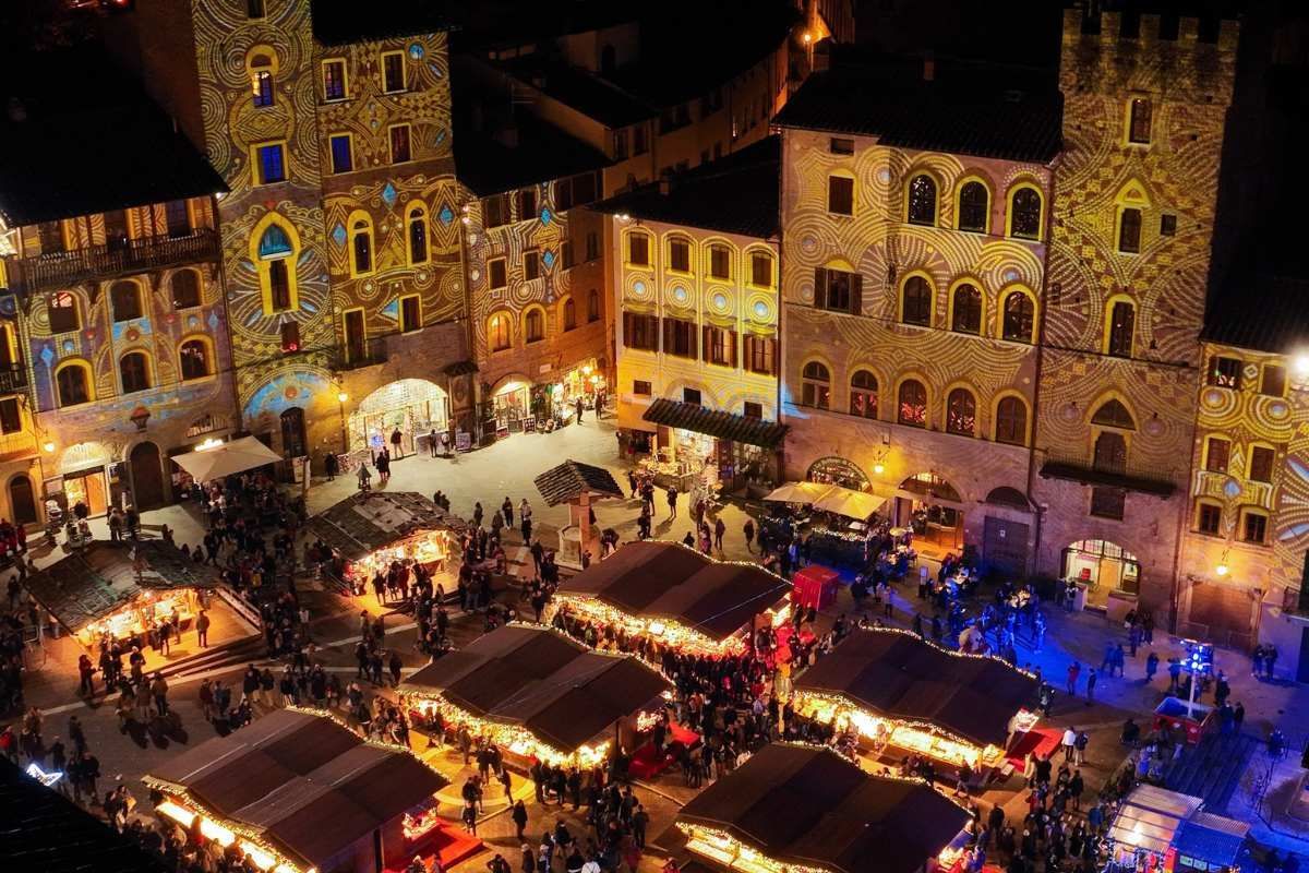 Una vista aerea di una piazza affollata di notte con bancarelle del mercato ed edifici illuminati da proiezioni di luce colorate.