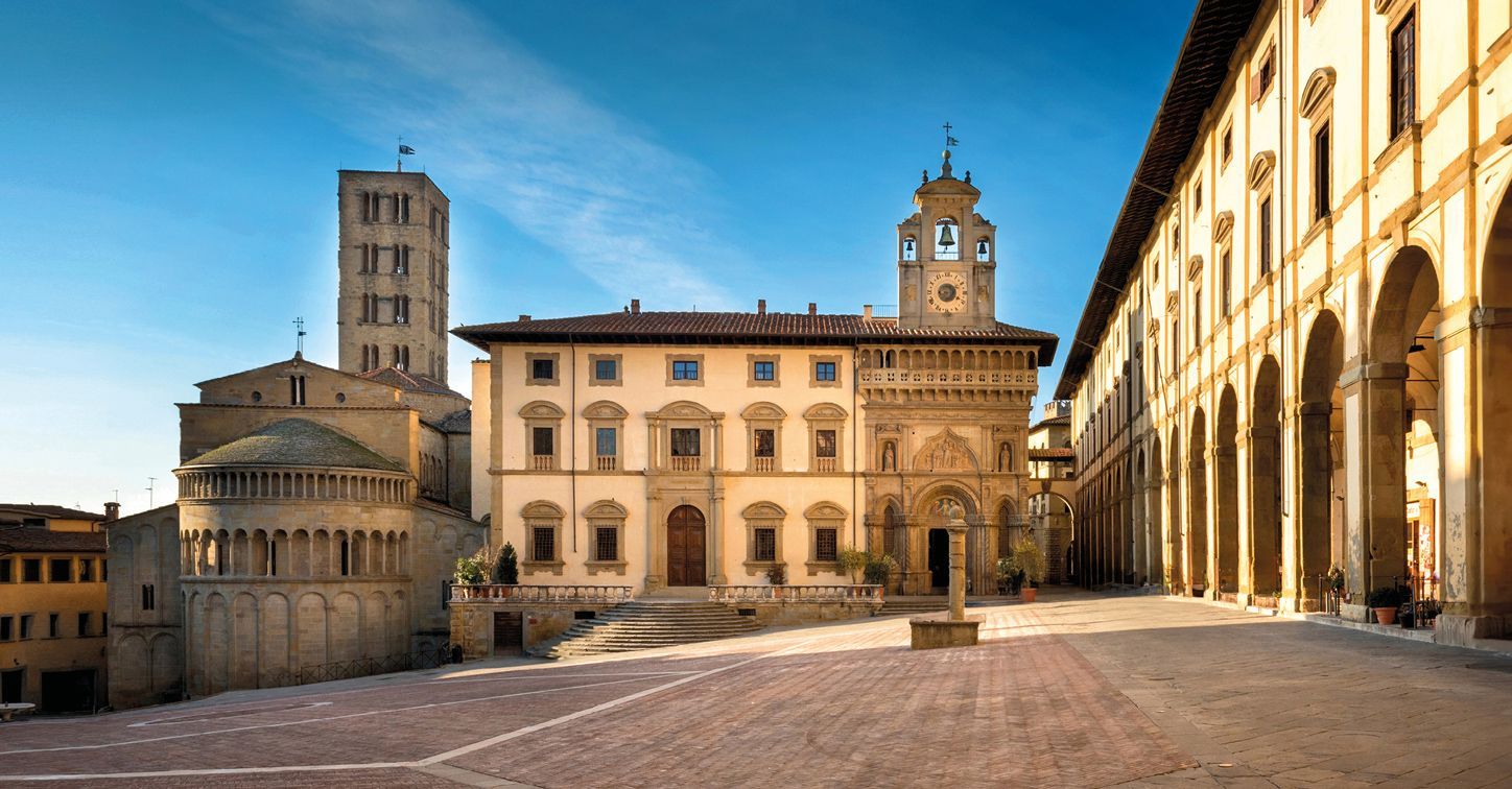 Una piazza in pietra vuota e illuminata dal sole, circondata da edifici storici, tra cui una chiesa, un campanile e una loggia ad archi, sotto un cielo azzurro.