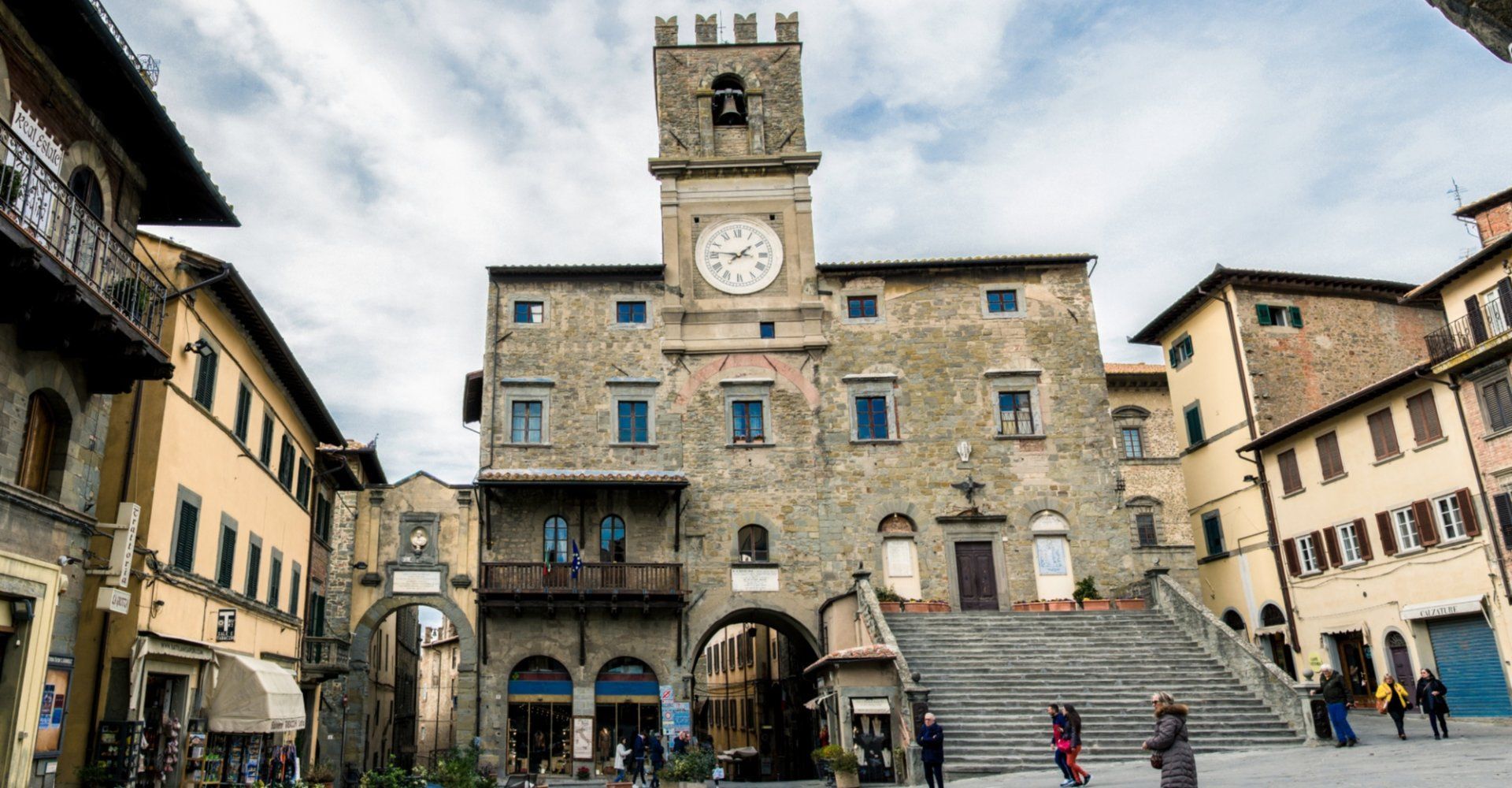 Un edificio storico in pietra con una grande torre dell'orologio e ampie scale si erge in una piazza europea sotto un cielo nuvoloso.