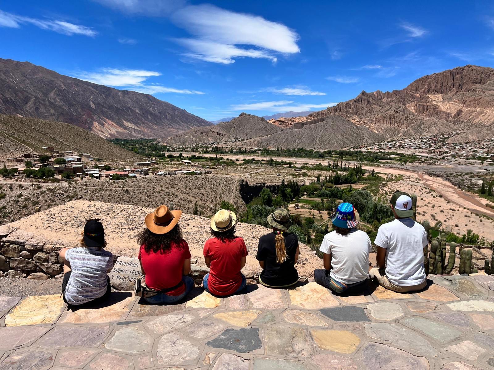 A WeRoad group trip sits with their backs to the camera on a stone viewpoint, looking out at a village in a vast mountain valley.