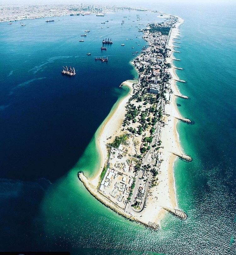 Vista aerea di una lunga penisola con spiagge, che separa una baia di un blu intenso, piena di navi, da acque costiere turchesi.