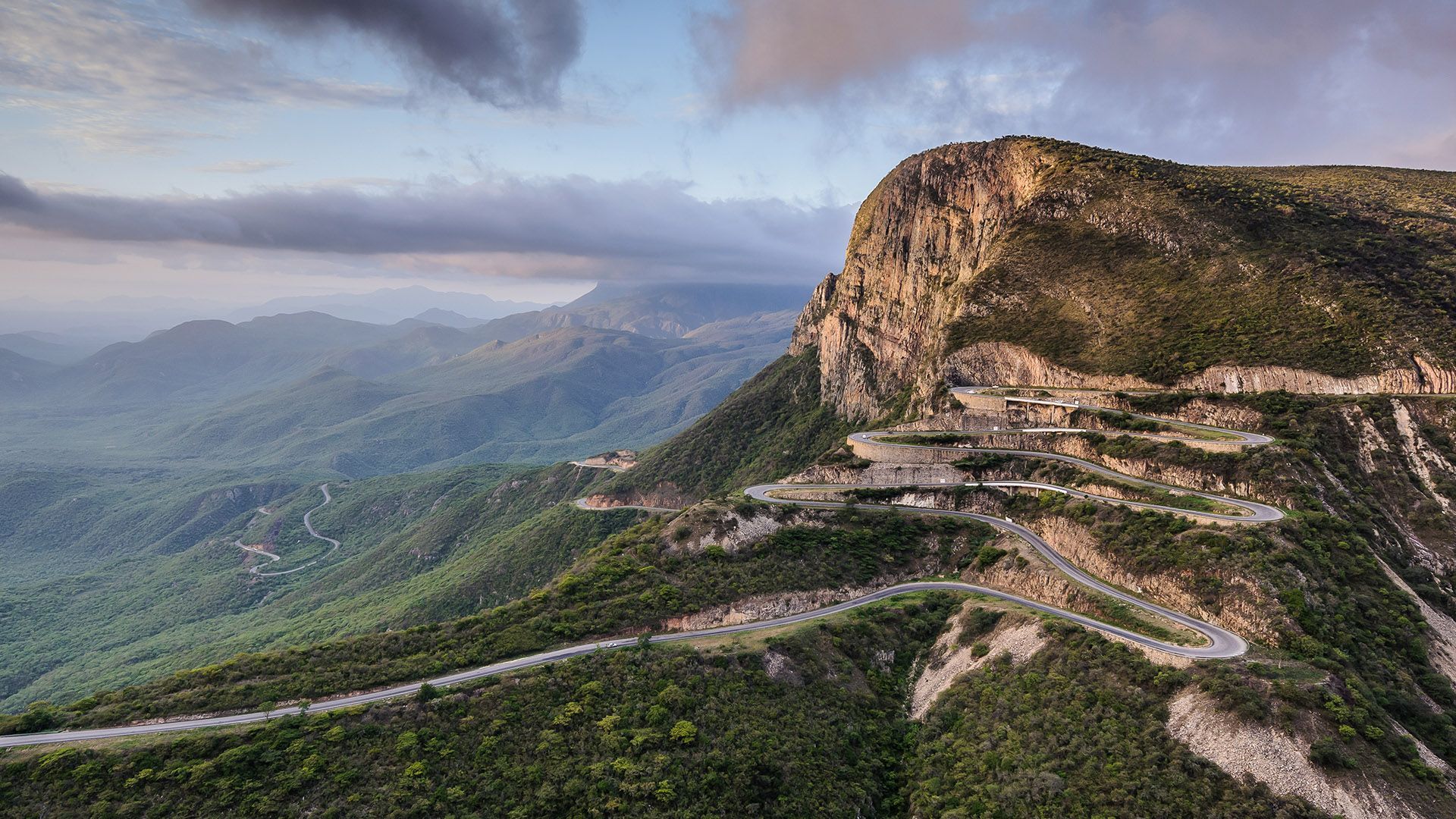 Vista aerea di una strada sinuosa con numerosi tornanti, scolpita in una montagna ripida e verdeggiante sotto un cielo nuvoloso.