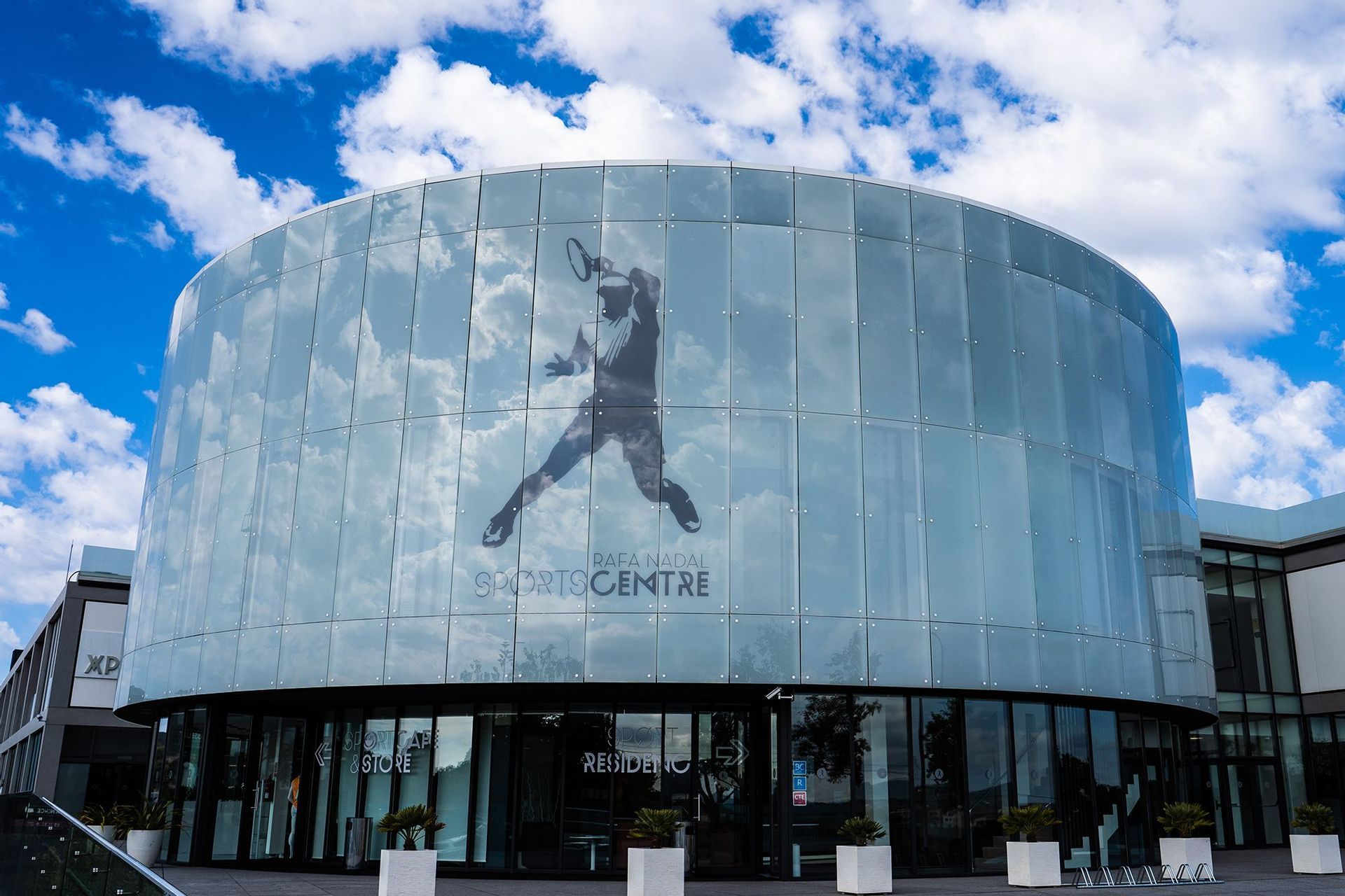 La facciata in vetro curvo dell'edificio Rafa Nadal Sports Centre, con una silhouette di un tennista sotto un cielo azzurro nuvoloso.