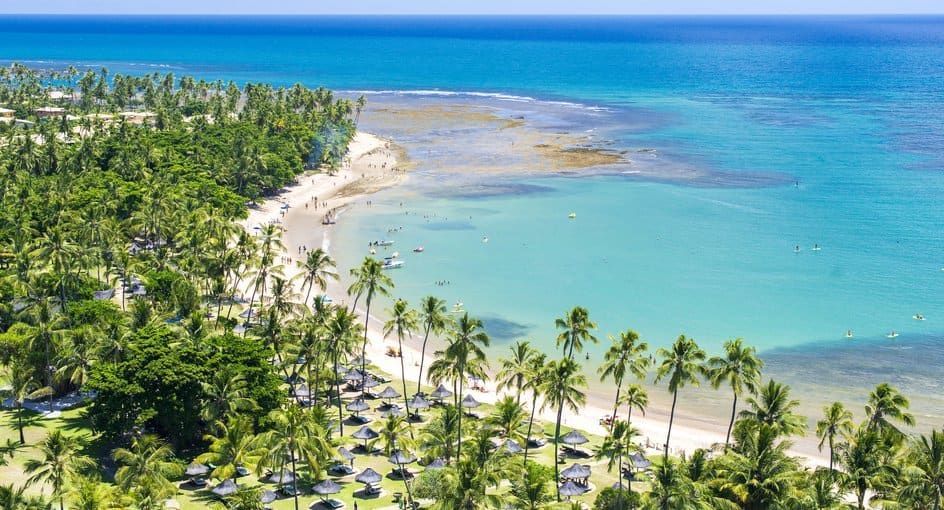 Vue aérienne d'une plage tropicale avec palmiers, parasols en chaume et personnes nageant dans l'océan turquoise.